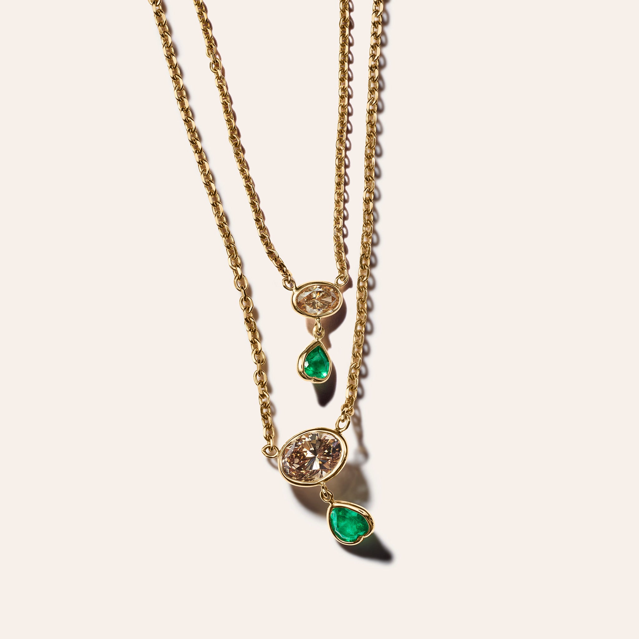 Two 18kt yellow gold chain necklaces layered together, each with a bezel-set east-west brown oval diamond and a dangling heart-shaped emerald charm in two sizes, shown against a cream background.
