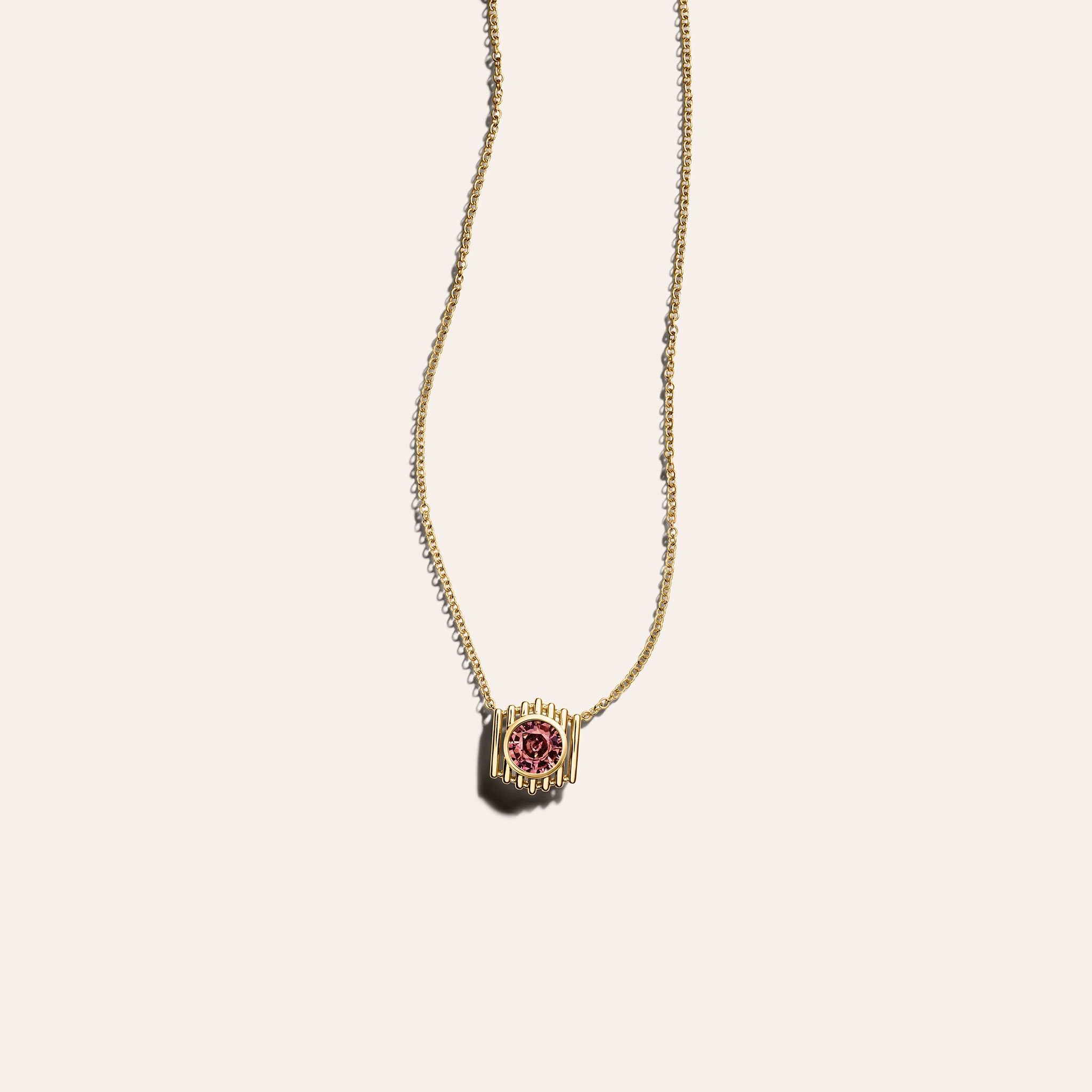 The Petite Prism Necklace in 18K yellow gold, with a bezel-set round peach sapphire framed by fluted gold bars on a fine cable chain, shown against a cream background.