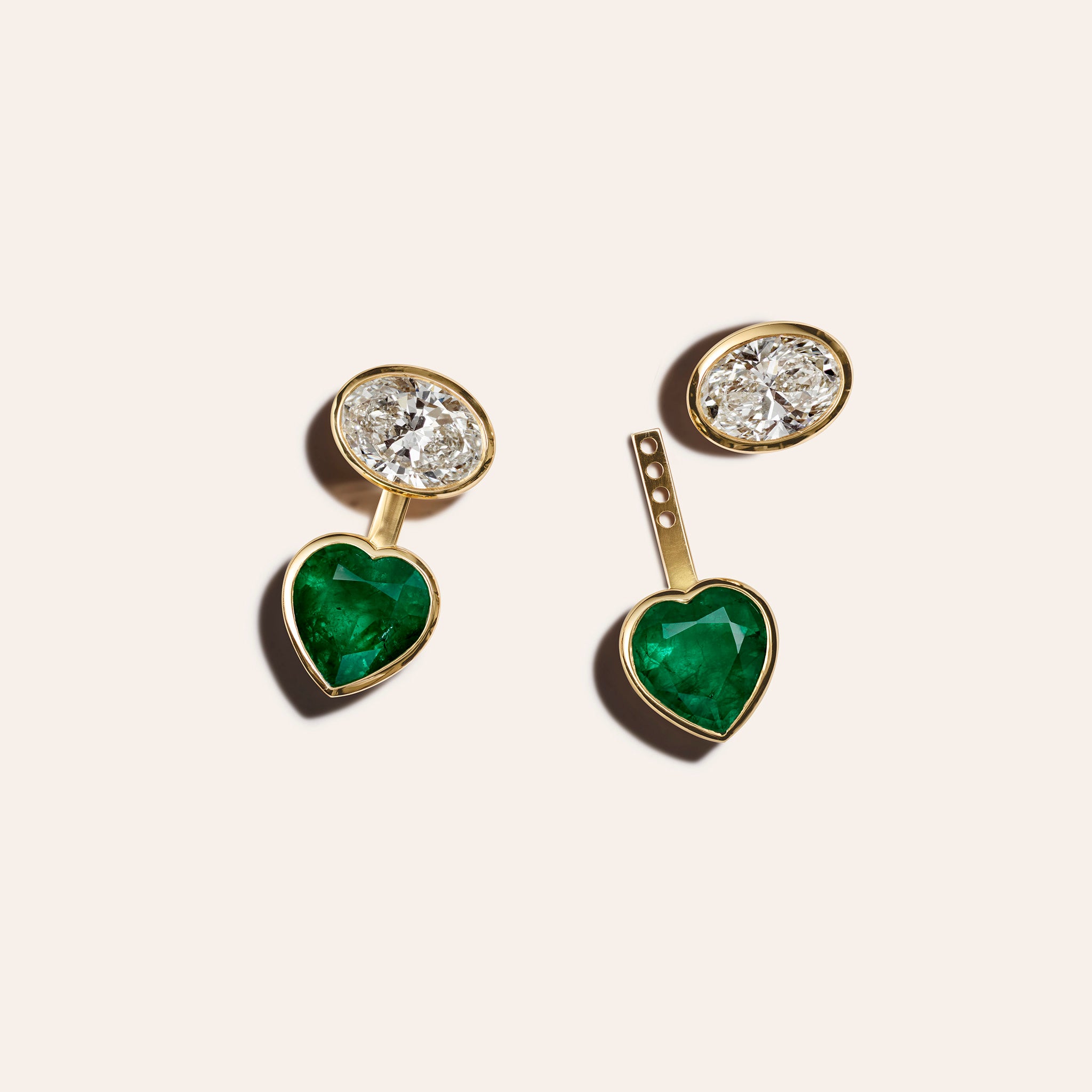 18kt yellow gold stud earrings with bezel-set white oval diamonds and removable heart-shaped emerald jackets, shown assembled and disassembled against a cream background.