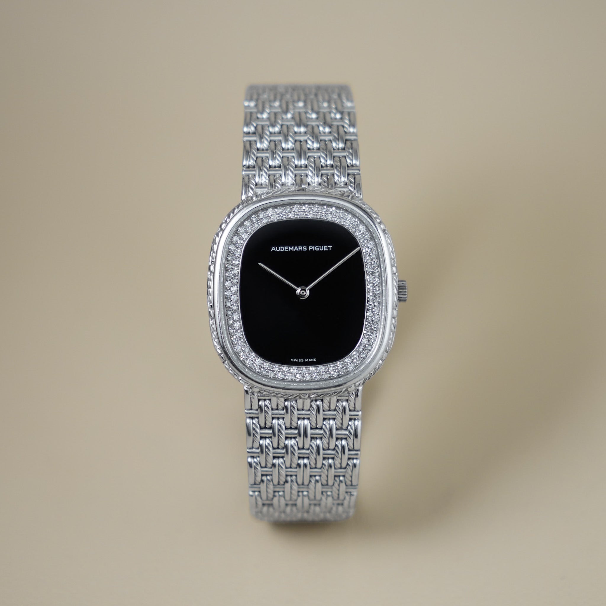 Audemars Piguet watch with a silver pavé bezel, silver woven mesh bracelet, and a black dial, centered on a warm beige background.