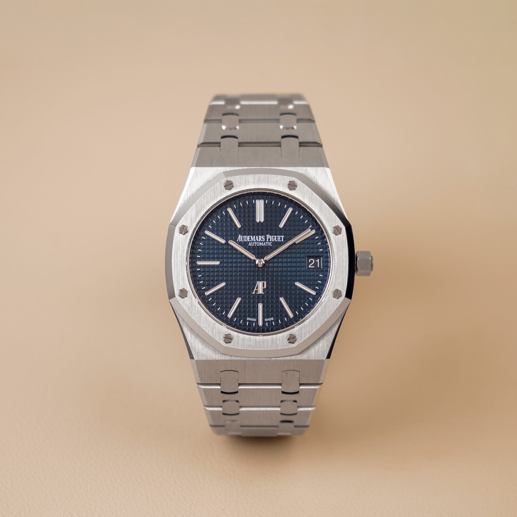 A steel Audemars Piguet Royal Oak with a steel bezel, textured blue dial with a date window, and steel bracelet, shot upright against a solid beige background.
