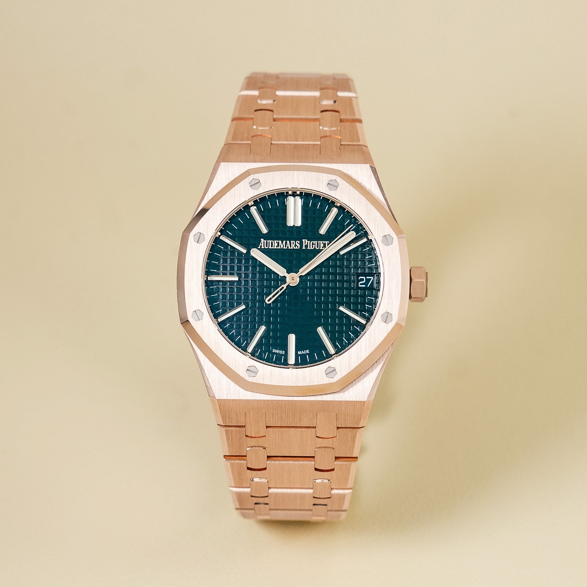 A rose gold Audemars Piguet watch with a textured black dial, an octagonal bezel, and a date window showing 27, shot against a neutral beige background.