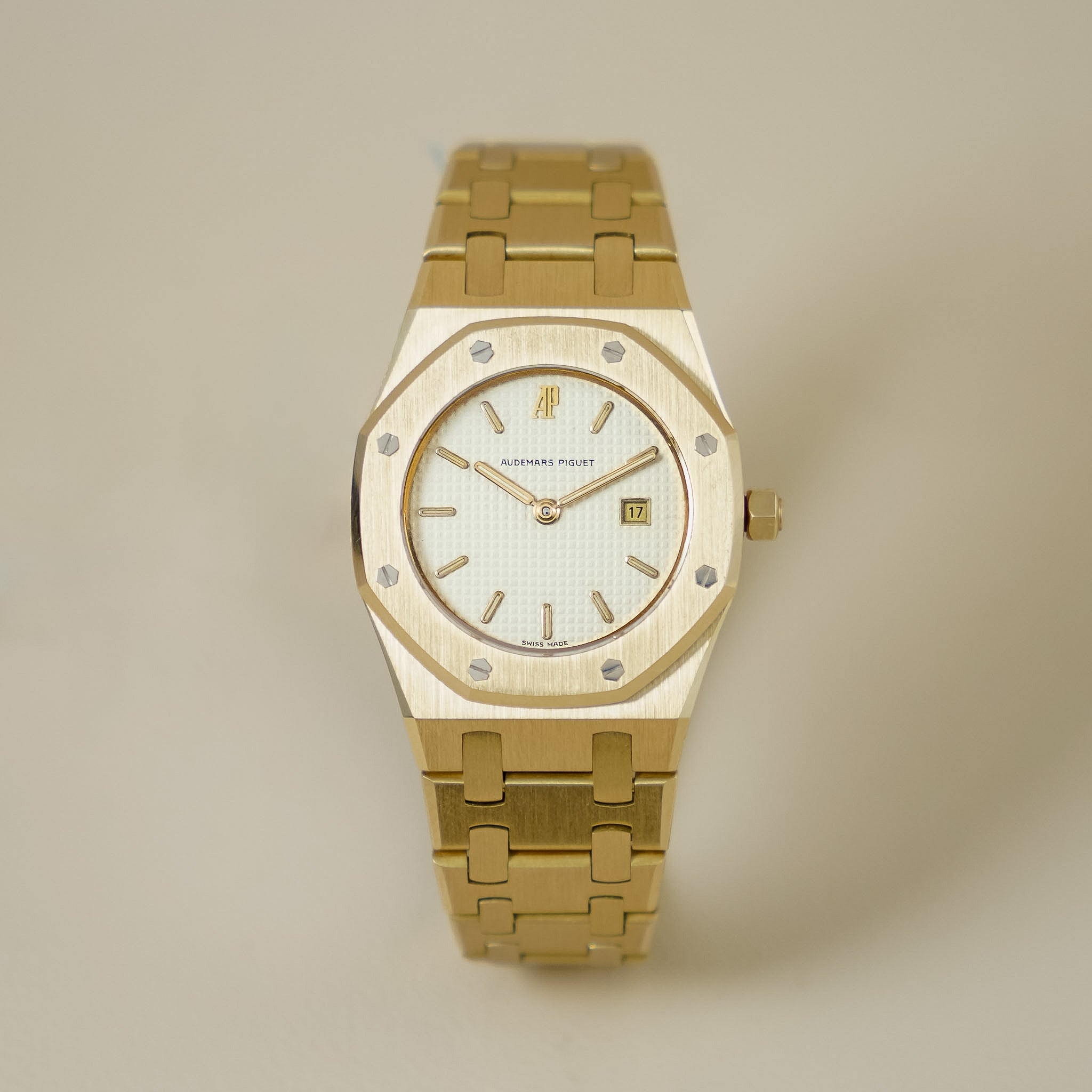A gold-tone wristwatch with an octagonal bezel, matching gold-tone link bracelet, and a white dial with gold indices, photographed against a solid light beige background.