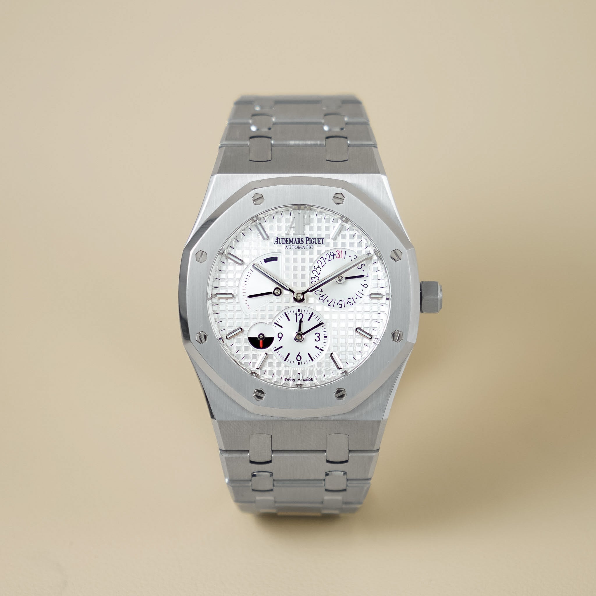 A silver stainless steel wristwatch with a white textured dial, featuring the Audemars Piguet logo on the face, set against a solid beige background.