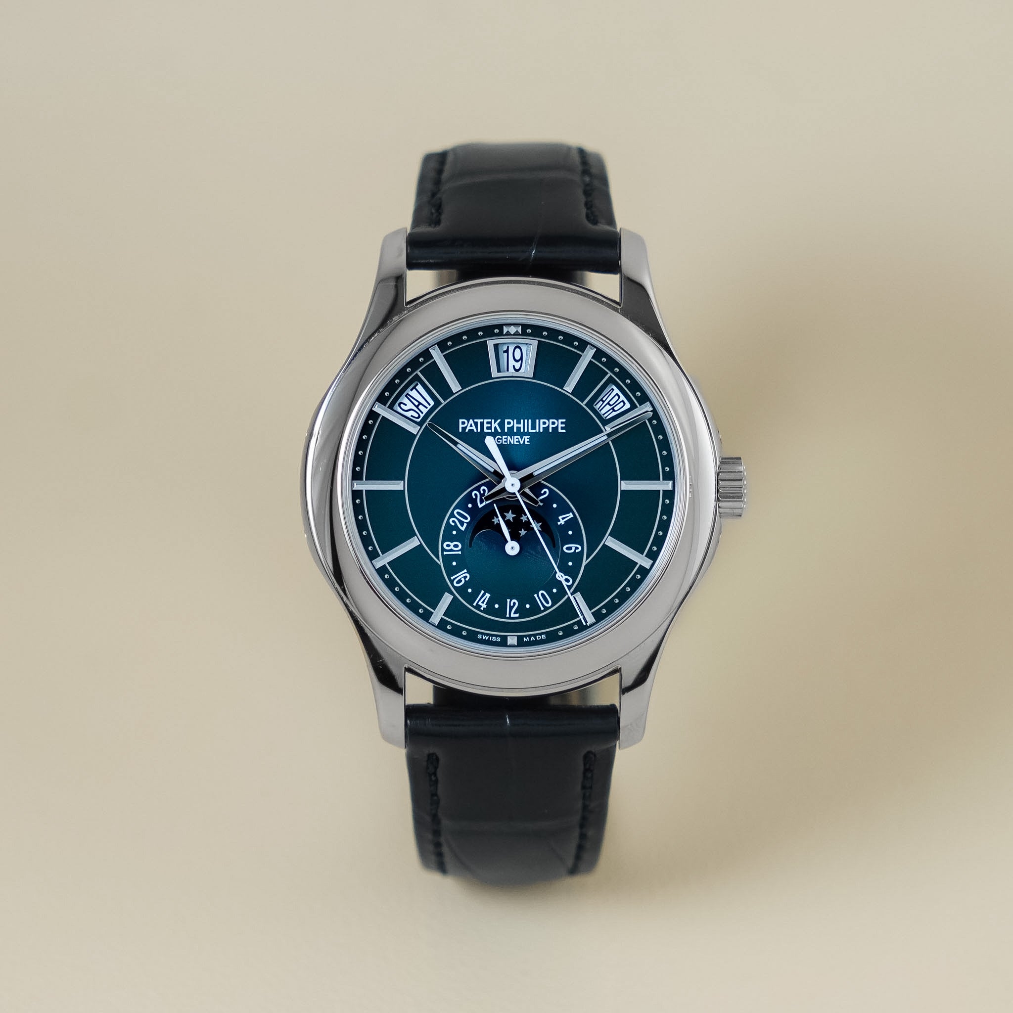 A Patek Philippe wristwatch with a silver-toned case, a dark teal blue dial, and a black leather strap, centered against a solid beige background.