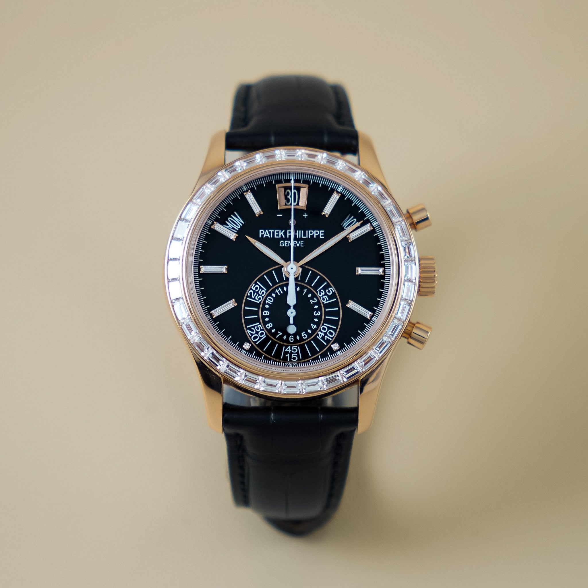 A rose gold chronograph watch with a black dial, diamond bezel, and black alligator strap against a neutral cream background