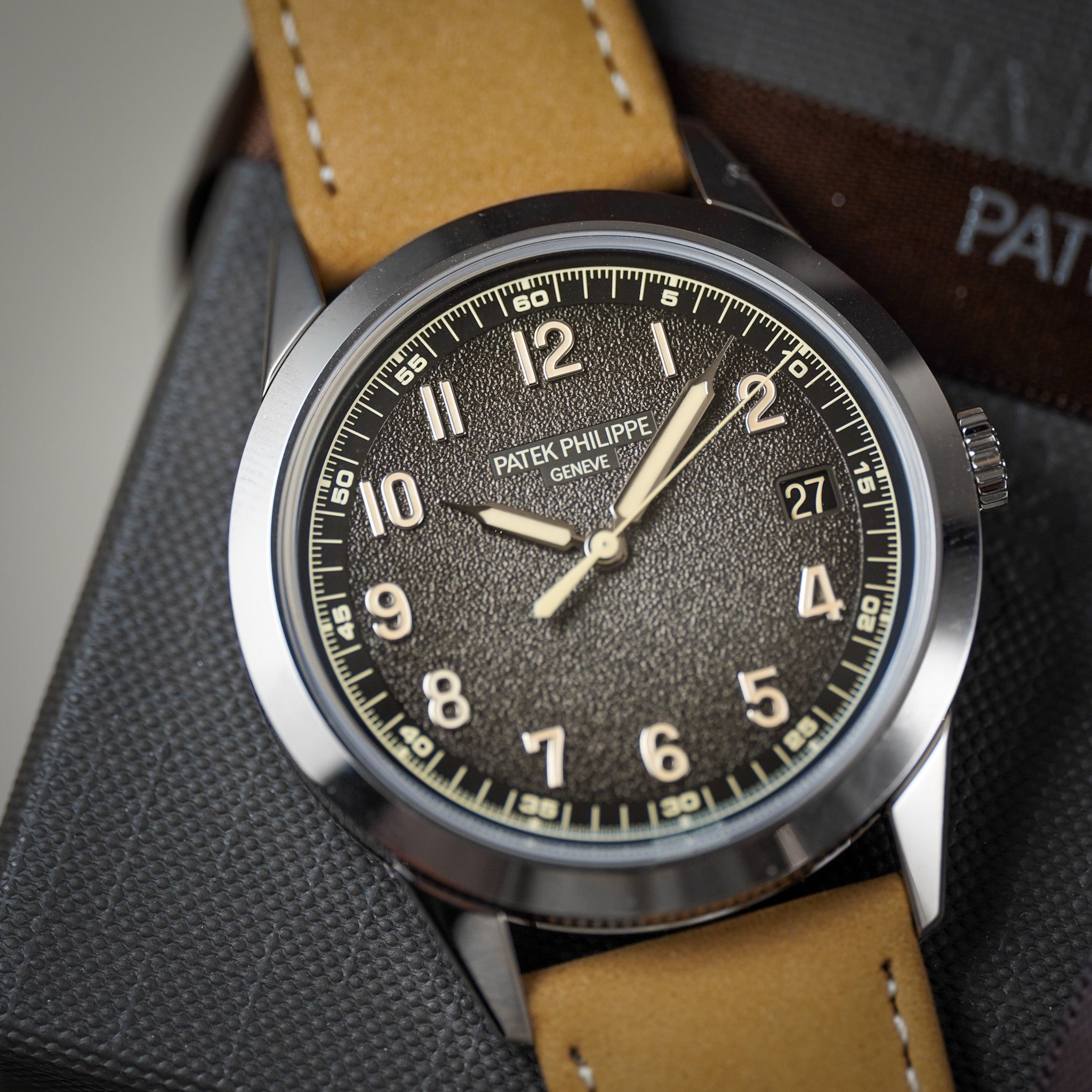 A military-inspired watch with a dark textured dial, tan leather strap, cream hour markers, laying on a grey booklet