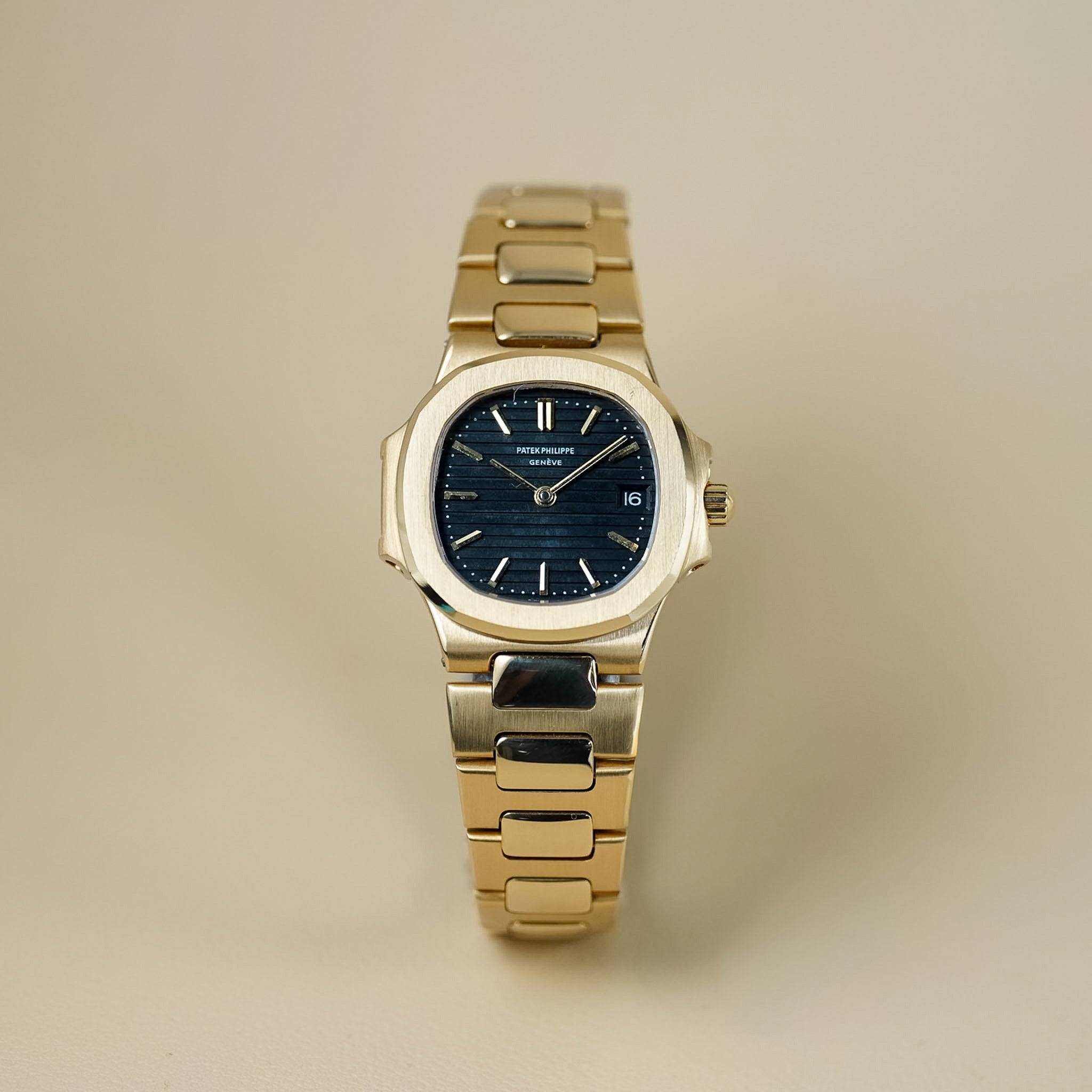 Patek Philippe ladies Nautilus watch with a yellow gold case and bracelet. The watch has a blue dial and is shot against a neutral beige background
