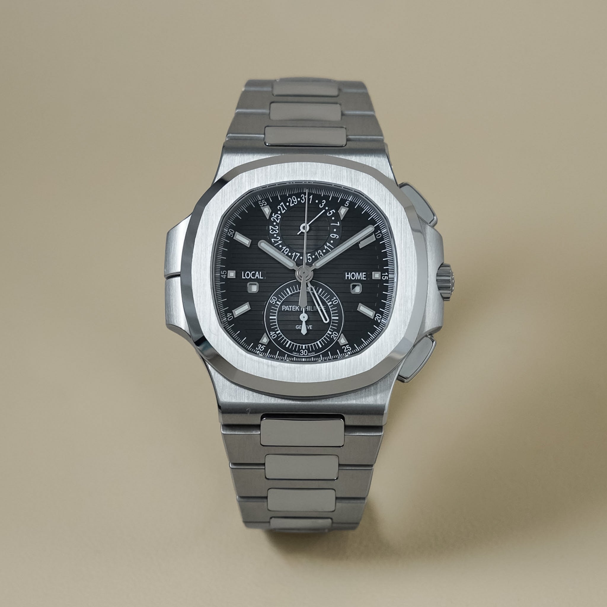 A stainless steel chronograph watch with a black dial and silver-toned metal bracelet, photographed from the front on a warm beige background.