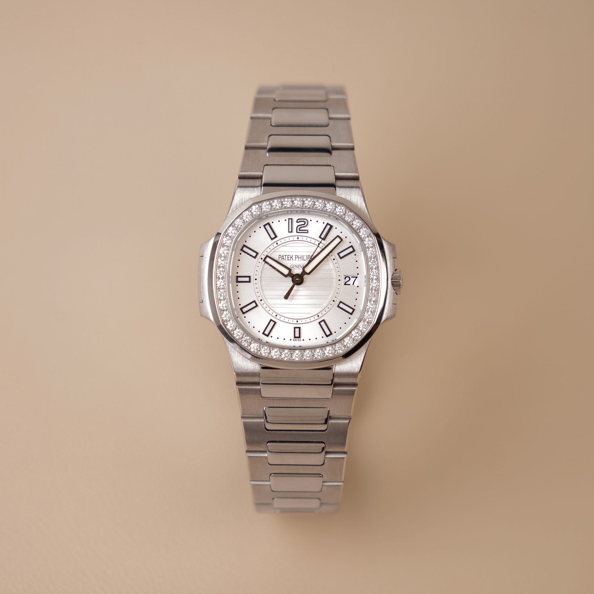 A white gold ladies Patek Philippe Nautilus with a white dial, diamond bezel, and white gold bracelet. Shot upright on a solid beige background. 