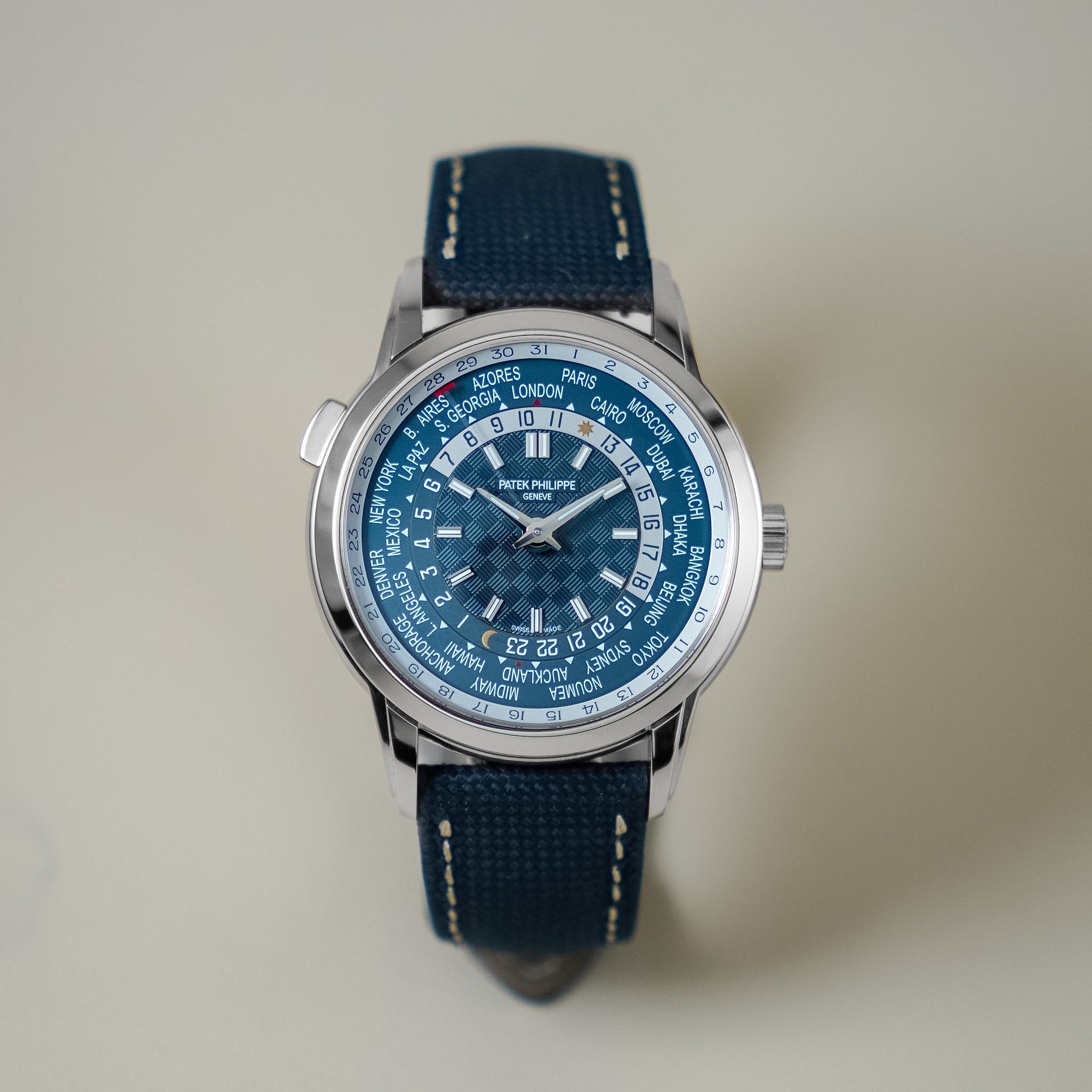 A wristwatch with a blue dial and a dark blue fabric strap, photographed against a plain light beige background. The watch case is silver-toned metal, and the dial features a world time layout with white hour markers and blue subdials.