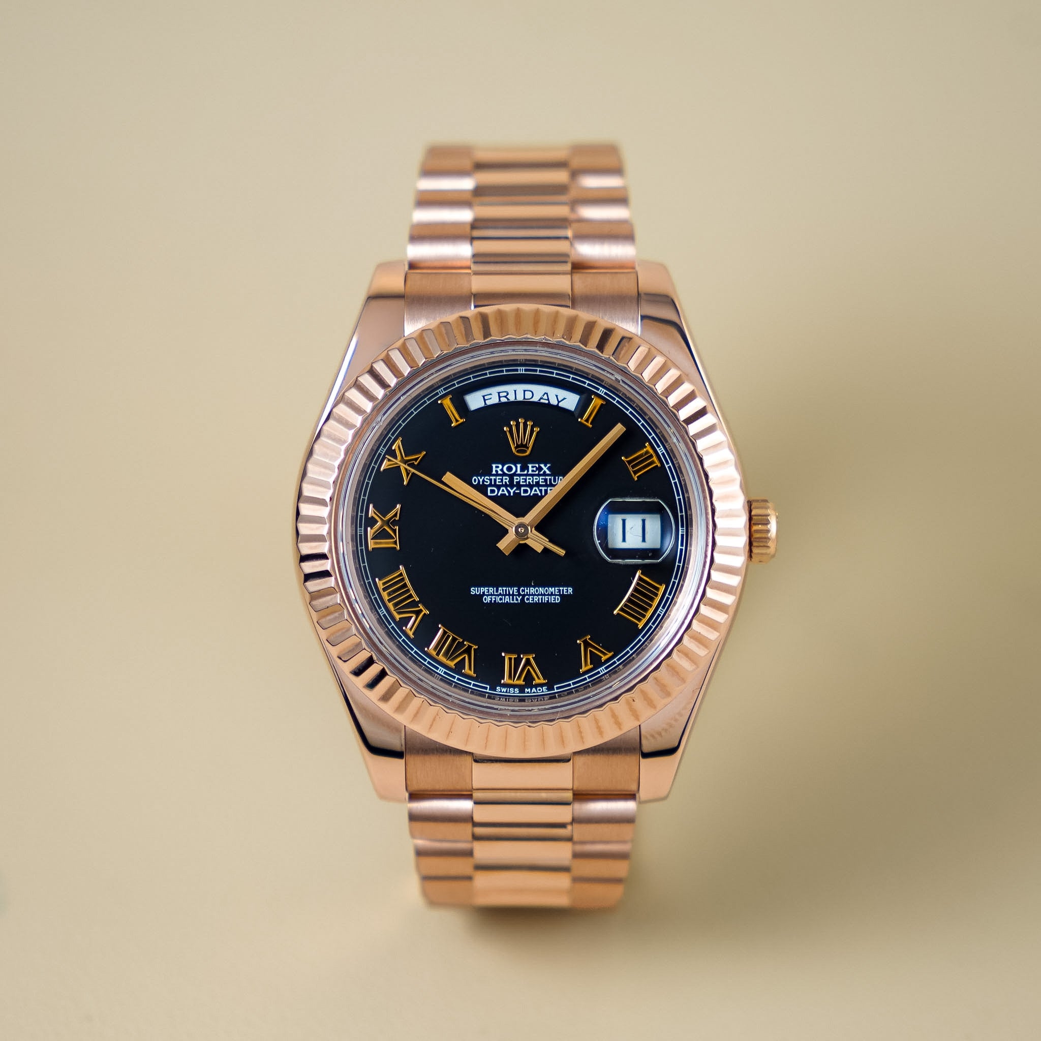 A head-on image of a rose gold Rolex Day-Date with a black dial and Roman numeral hour markers against a neutral cream background.