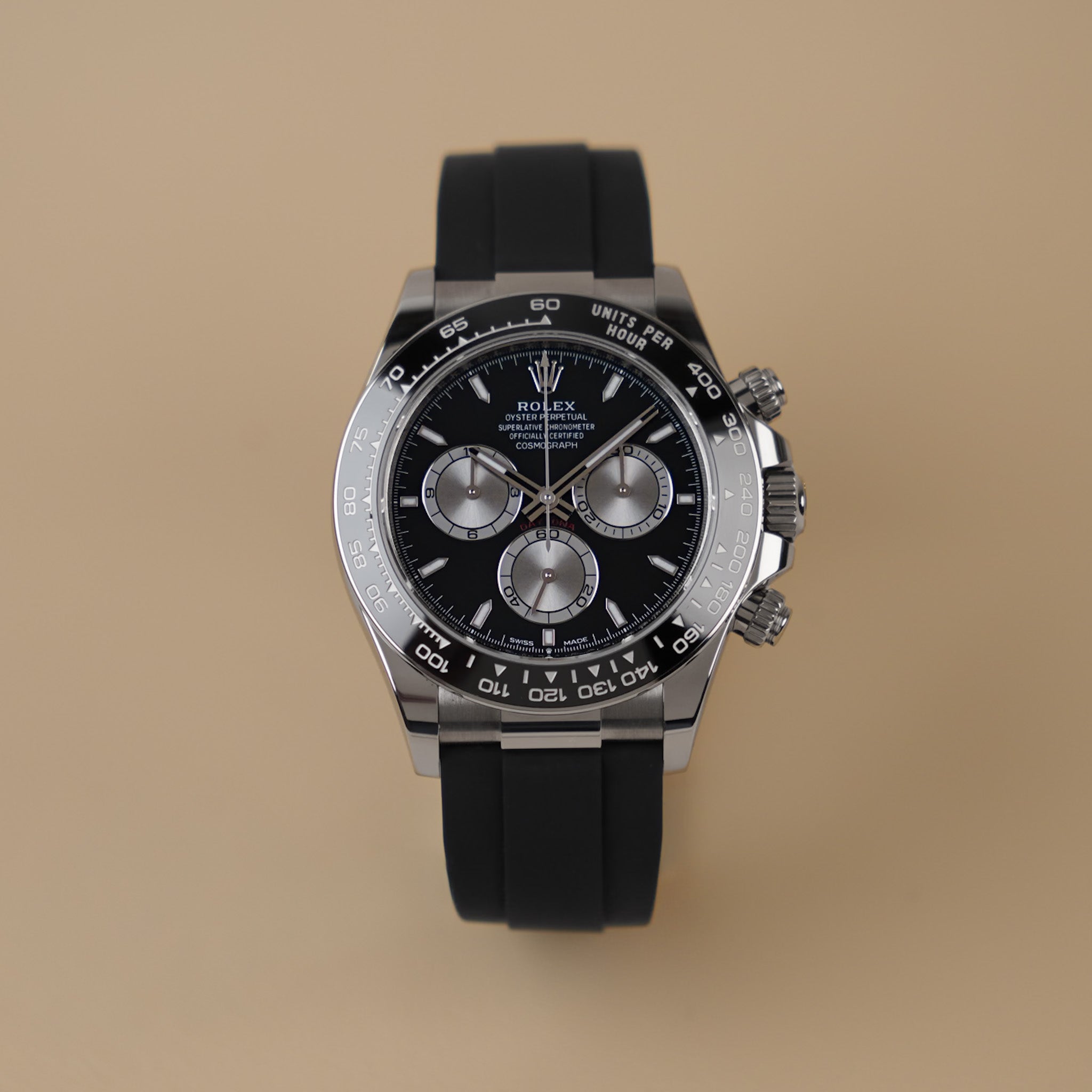 A Rolex Daytona in white gold with a black dial, silver subdials, a black ceramic bezel, and a black rubber strap. Shot upright on neutral beige background