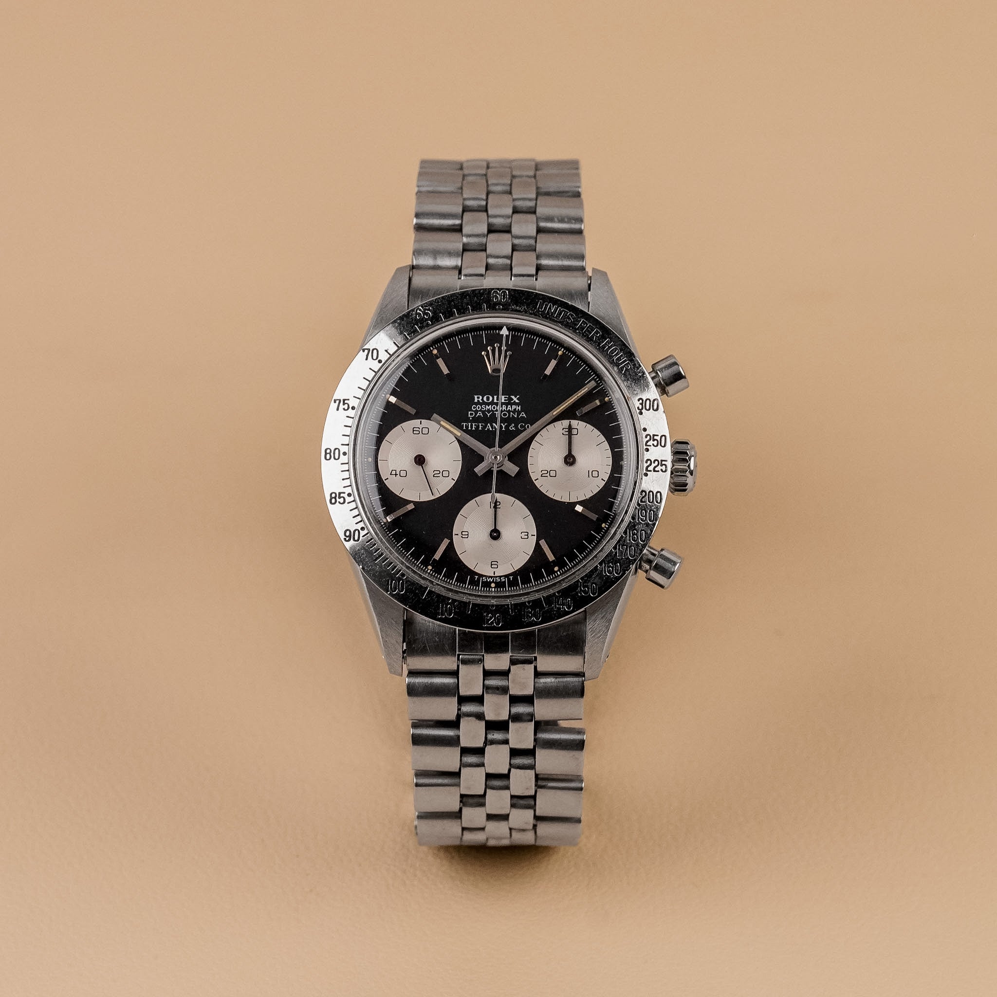 A steel vintage Rolex Daytona with a lack dial, white subdials, steel bezel, and steel bracelet, shot upright against a solid beige background