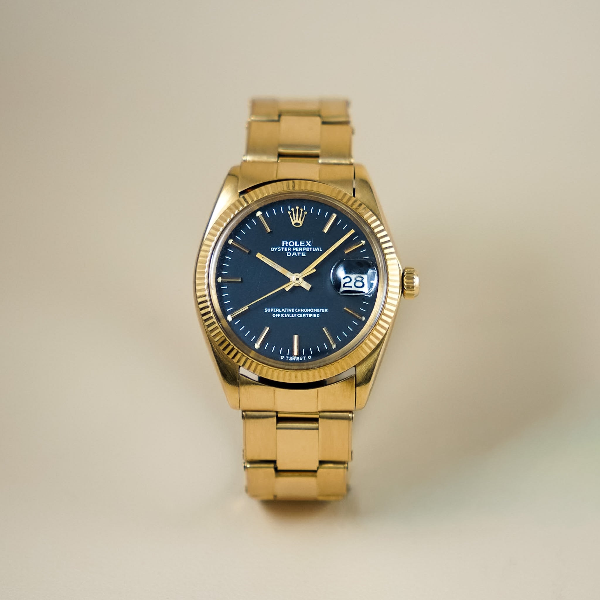 A yellow gold Rolex watch with a black dial, fluted bezel, and date window showing "28"