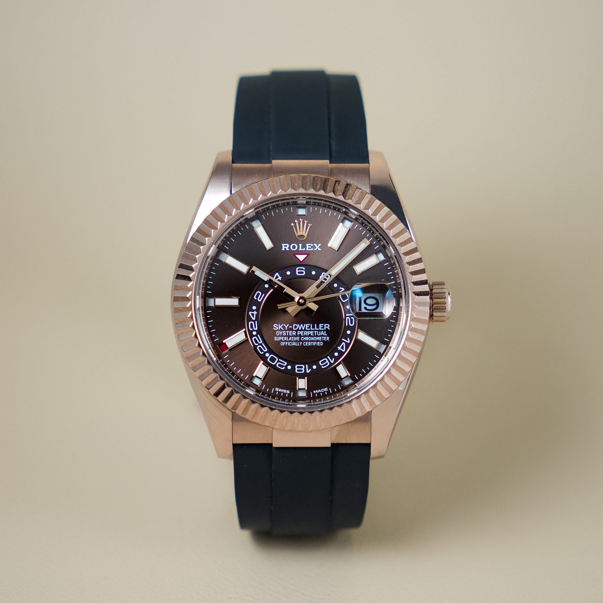 A head-on view of the rose gold Rolex Sky-Dweller watch with a chocolate brown dial on a black rubber strap against a neutral cream background