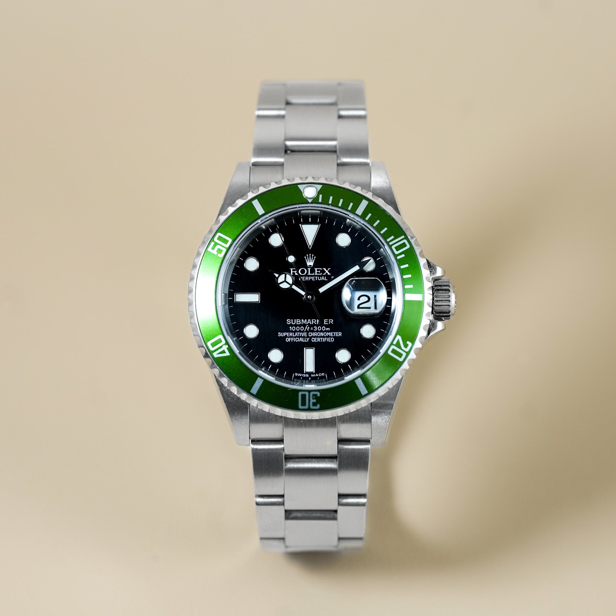 A Rolex Submariner Date watch with a stainless steel Oyster bracelet and a green aluminum bezel, shown against a solid beige background.