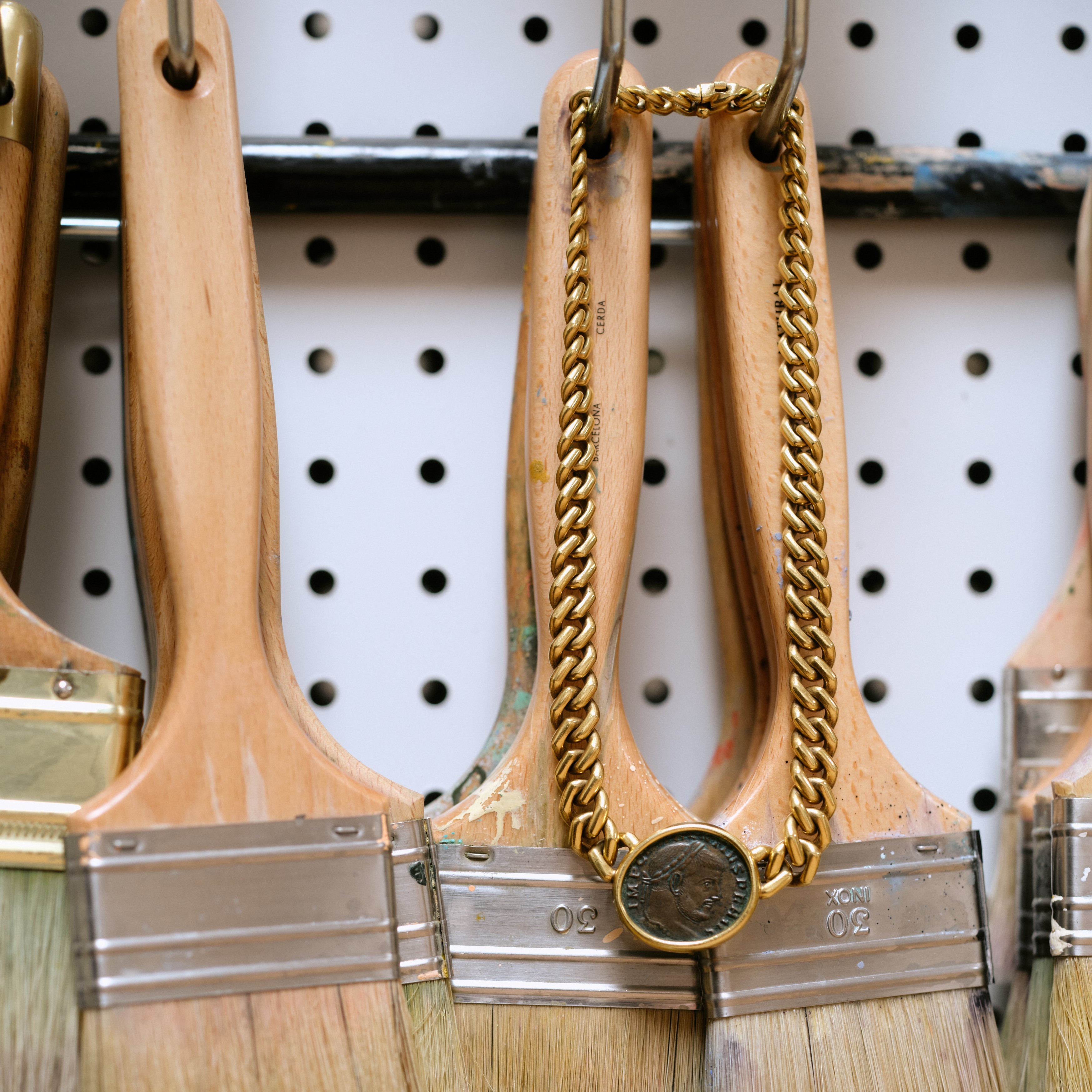 A close-up of hanging paint brushes with wooden handles and silver metal bristle caps, with one brush decorated by a gold chain draped over its handle and a round coin charm at the bottom, set against a white pegboard background.