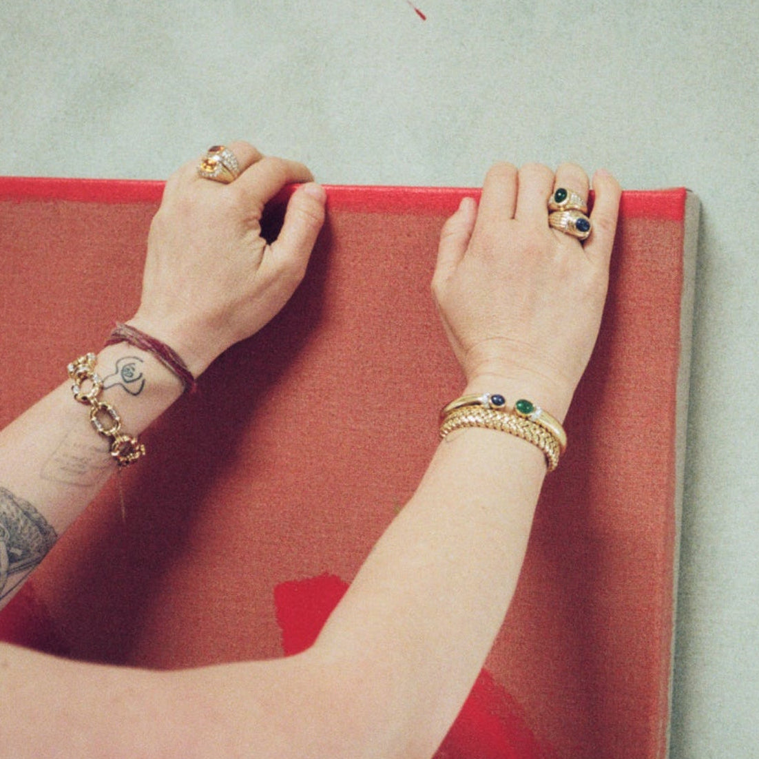 Hands wearing a vintage Boucheron lapis ring alongside layered gold rings and bracelets, pressing against a red painted canvas