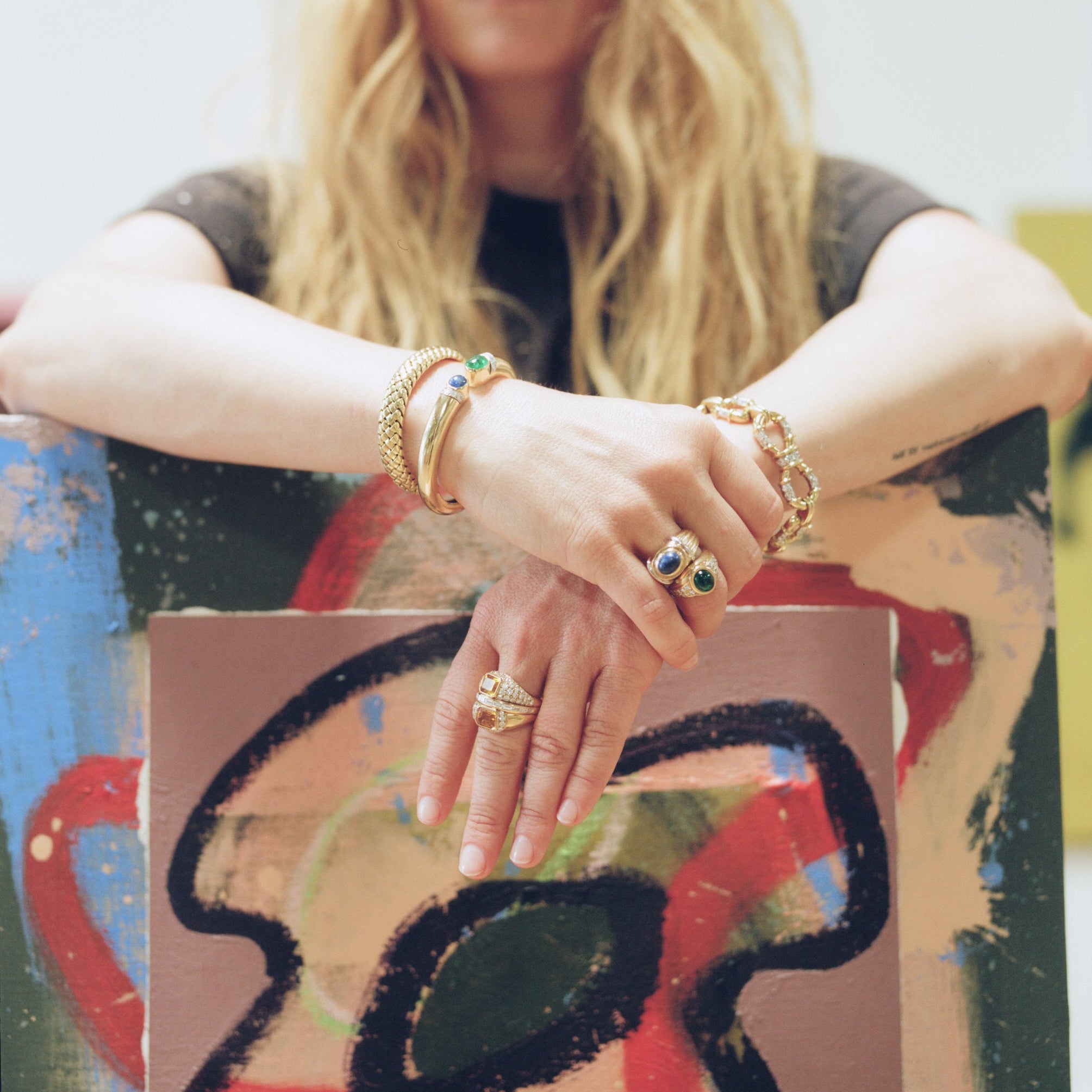 A woman with long blonde hair leans against a colorful abstract painting, wearing multiple gold rings and bracelets. The photo is softly lit with a blurred background and a large abstract artwork in the foreground.