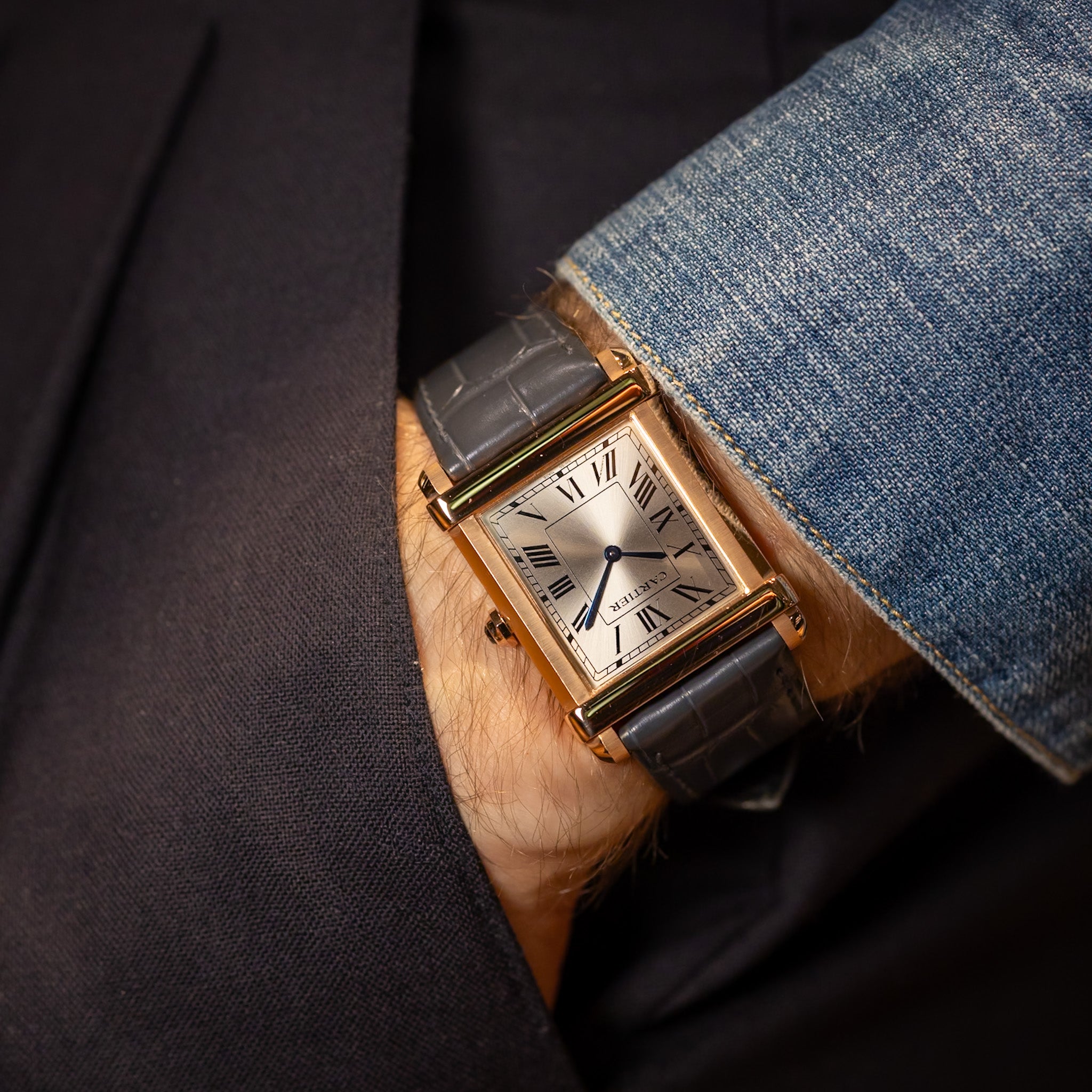 Luxury watch with brown leather strap on a person's wrist, wearing a dark suit and blue jeans.