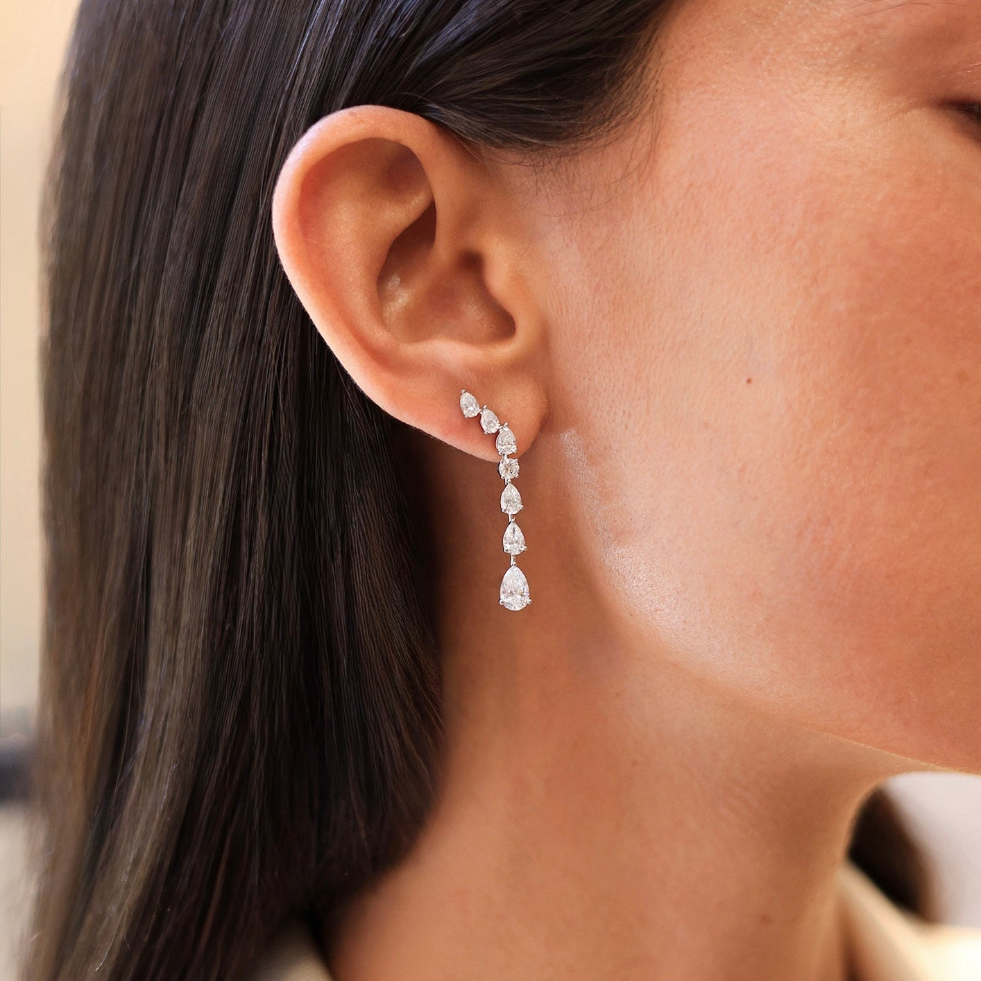 A close-up of a model's ear wearing a delicate curved drop earring set with small clear stones and a teardrop-shaped drop, shown against dark hair and a light neutral background.