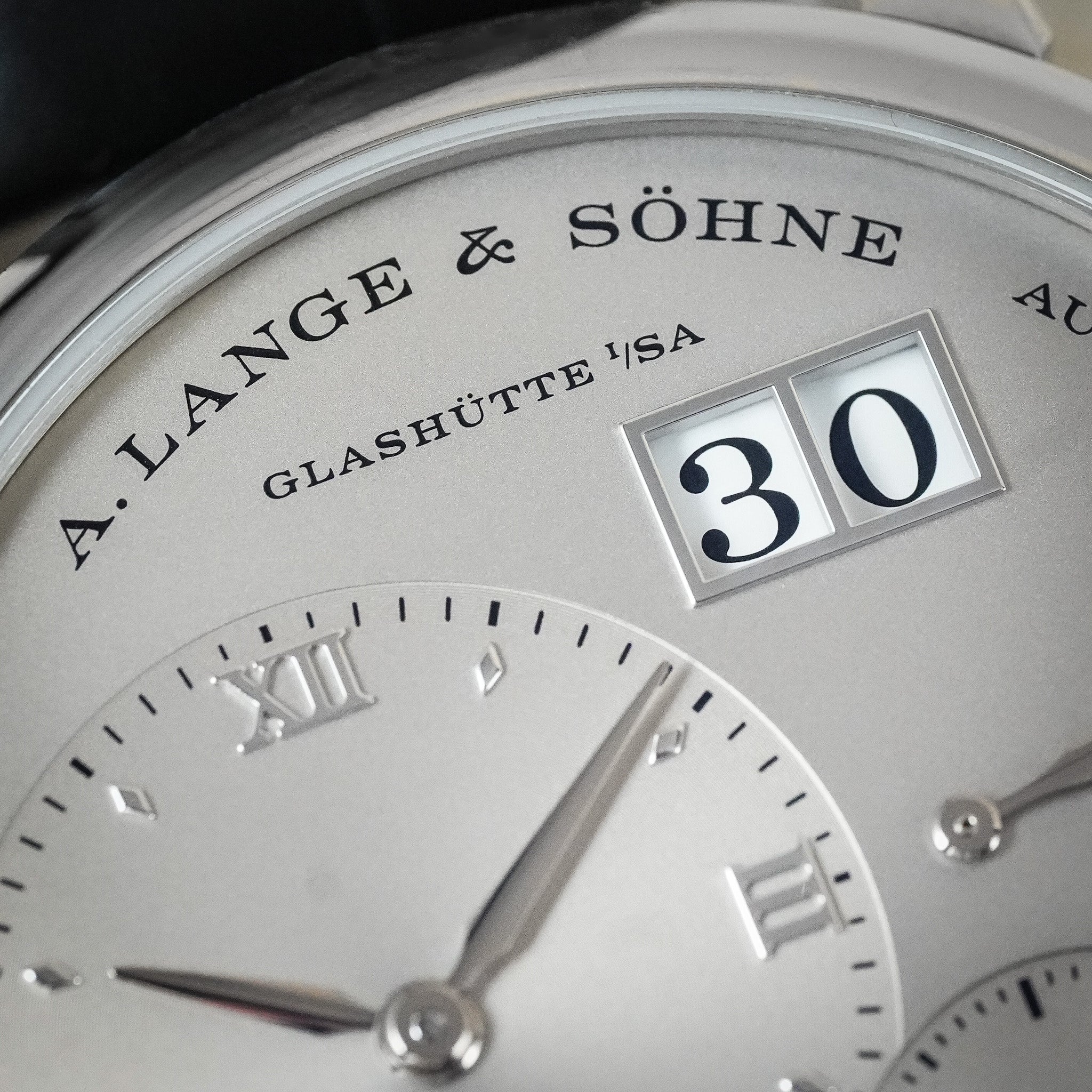 Close-up of a silver A. Lange & Söhne Glashütte 1'SA watch dial with Roman numeral hour markers, a date window showing 30, and polished metal hands.