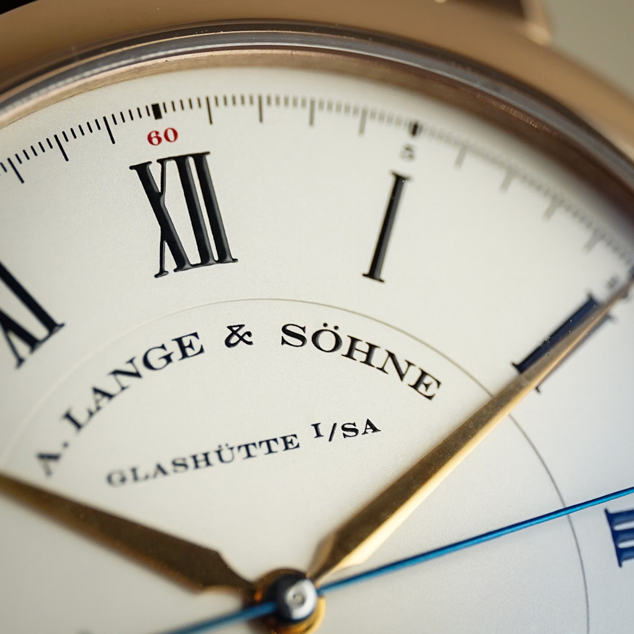 Close-up of a luxury watch dial with Roman numerals, gold hands, and the brand name A. Lange & Söhne Glashütte I/SA printed on an off-white face.