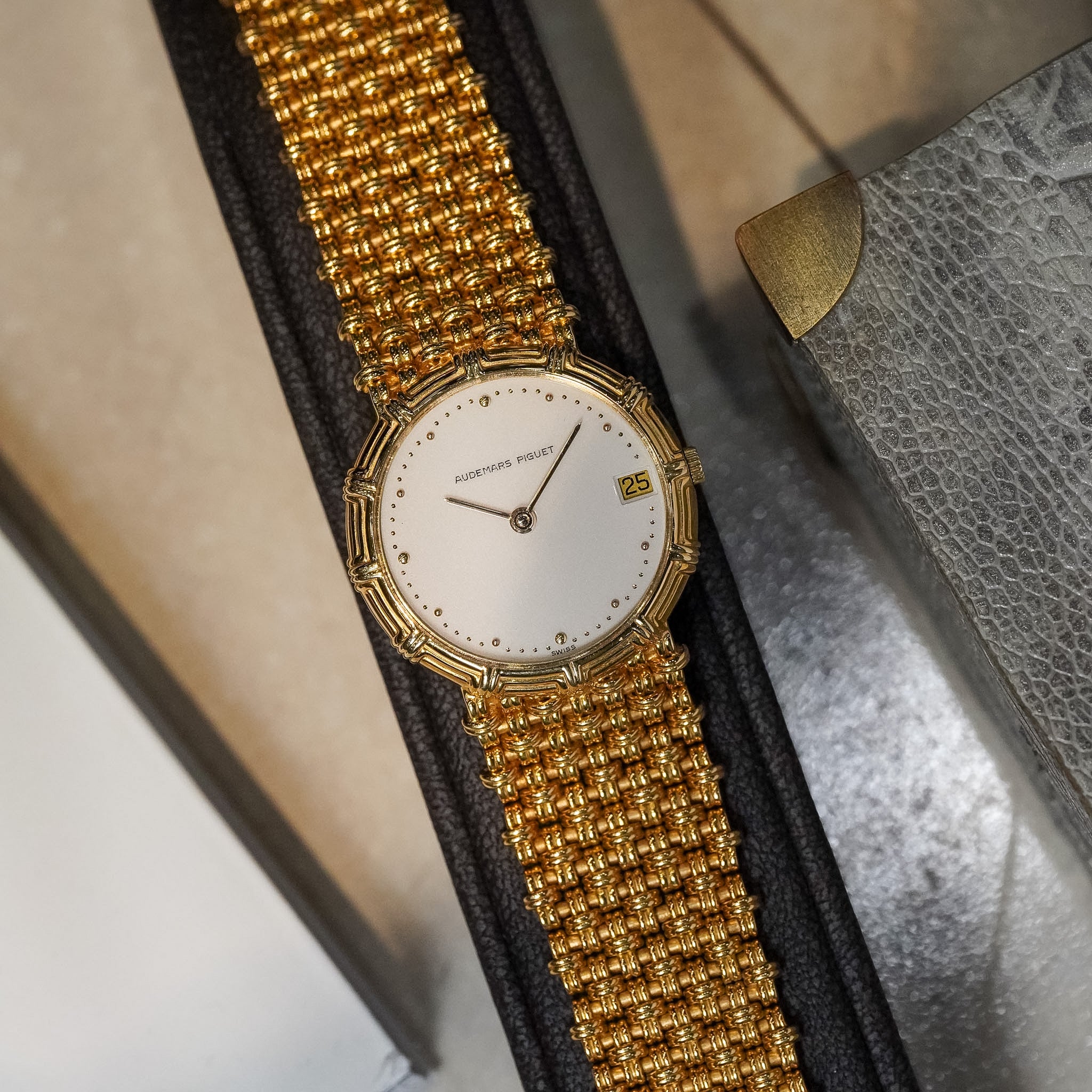A vintage-style wristwatch with a gold-toned beaded mesh strap and a white dial, resting on a black textured display surface next to a gray textured box.
