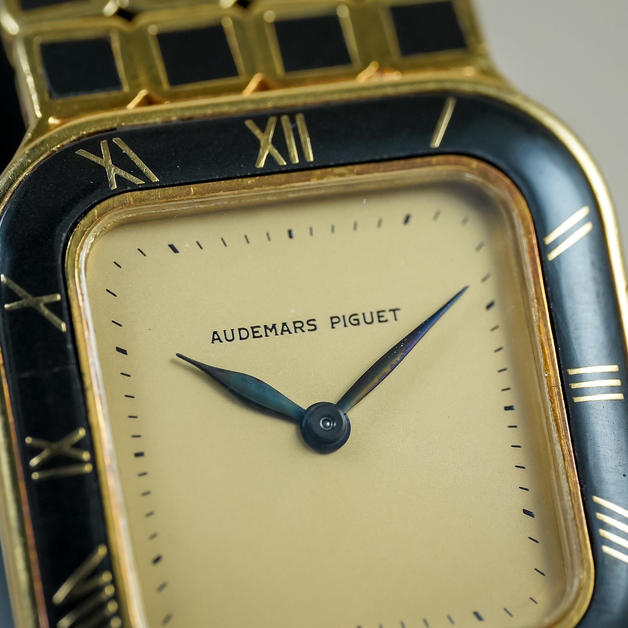 Close-up of an Audemars Piguet watch with a gold-tone case, black enamel hour markers, and a cream-colored dial with blue-toned hands.