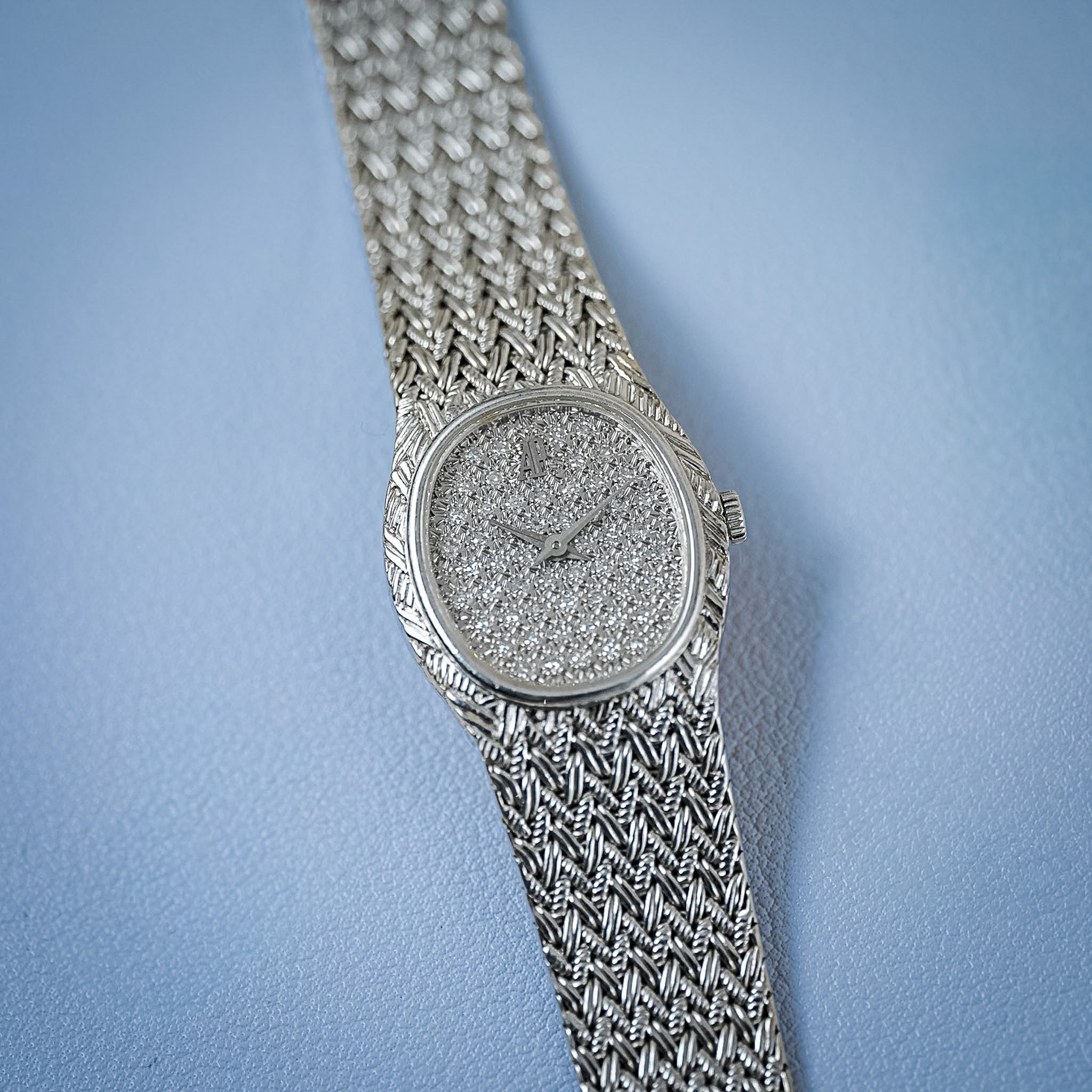 A white gold women's watch featuring a textured, diamond-encrusted dial and a matching knitted mesh bracelet, laying flat on a light blue surface