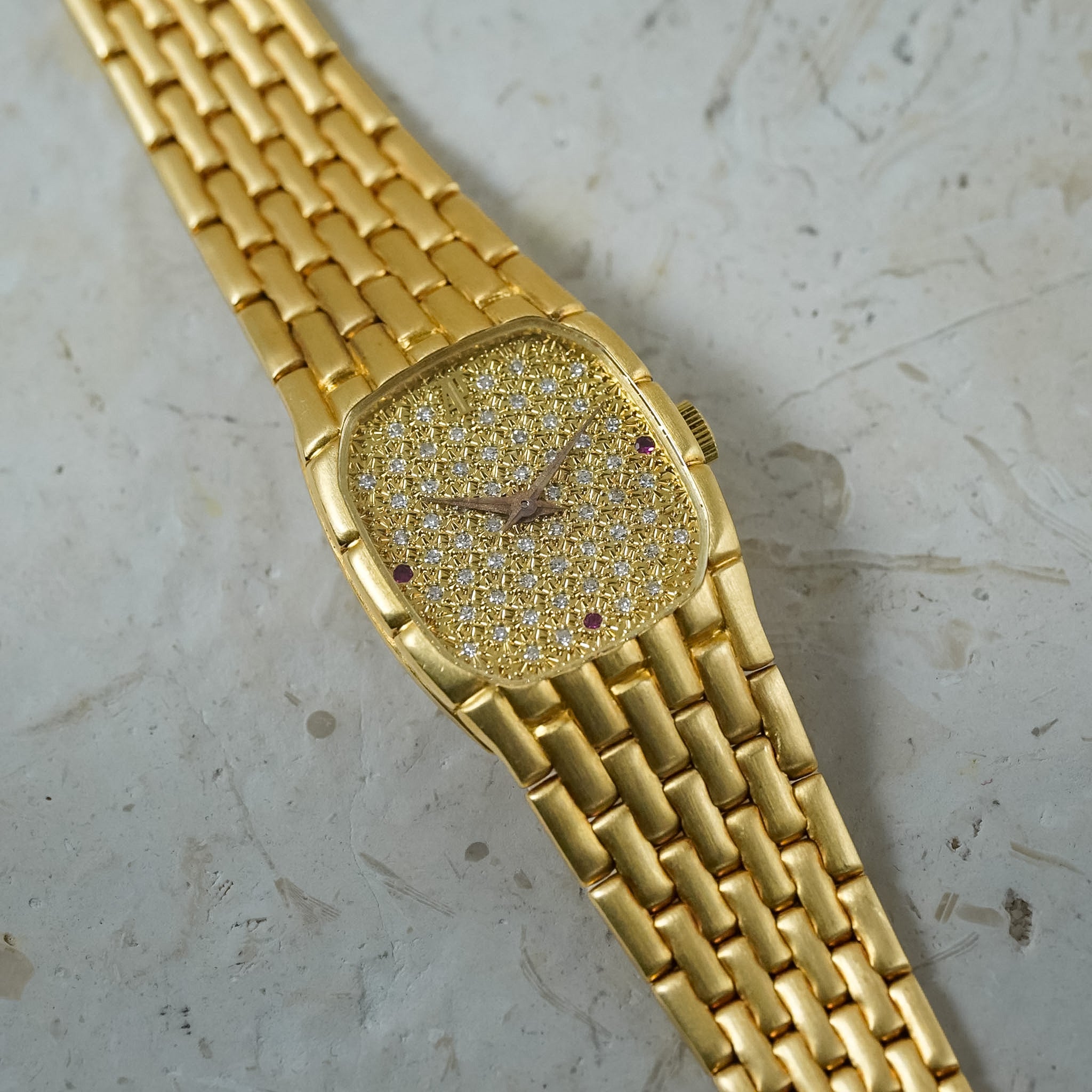 A gold watch with a pavé diamond dial and matching link bracelet, photographed diagonally on a textured light gray stone surface.