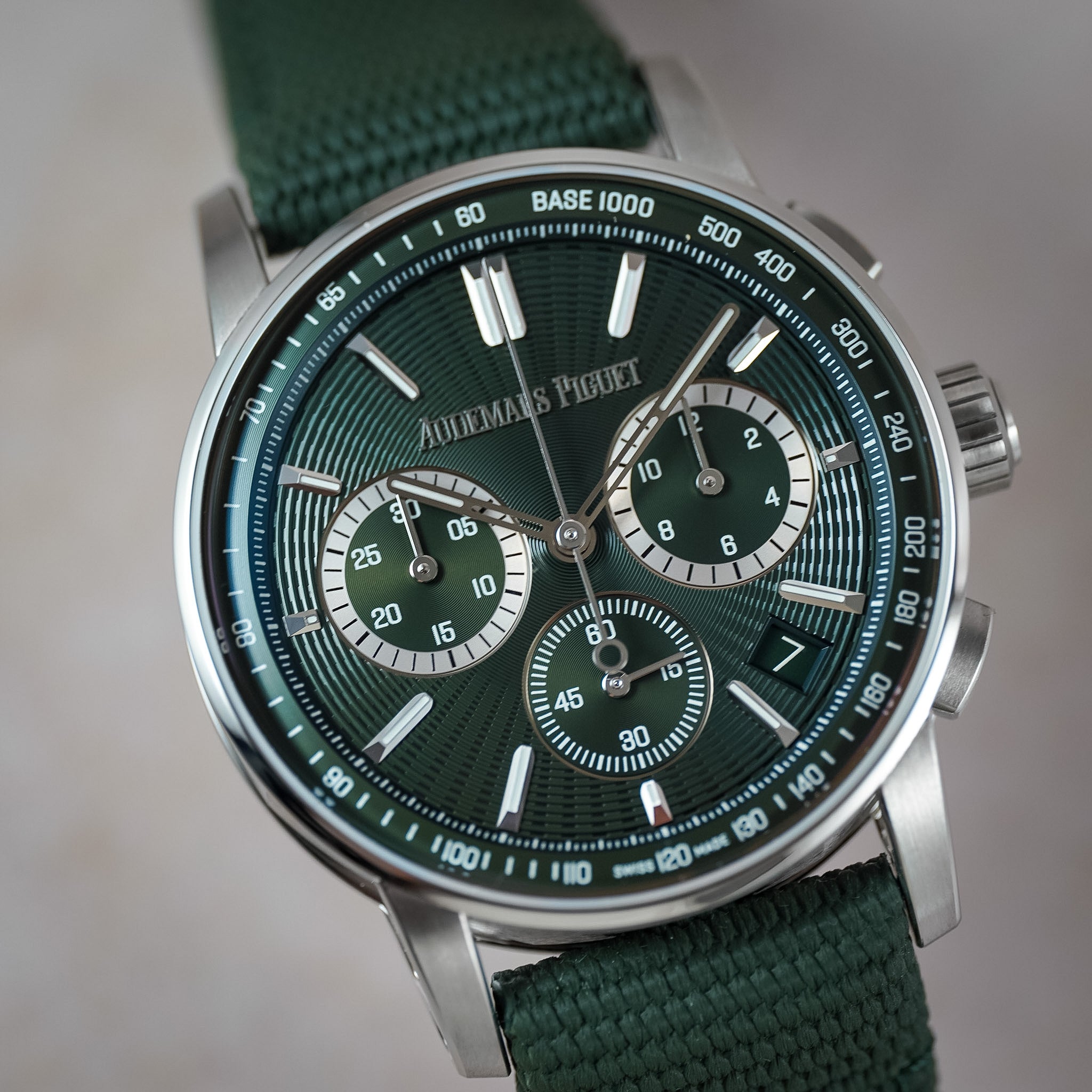 A close-up shot of a Audemars Piguet wristwatch featuring a dark green dial, a matching green fabric strap, and a silver stainless steel case with a tachymeter bezel.