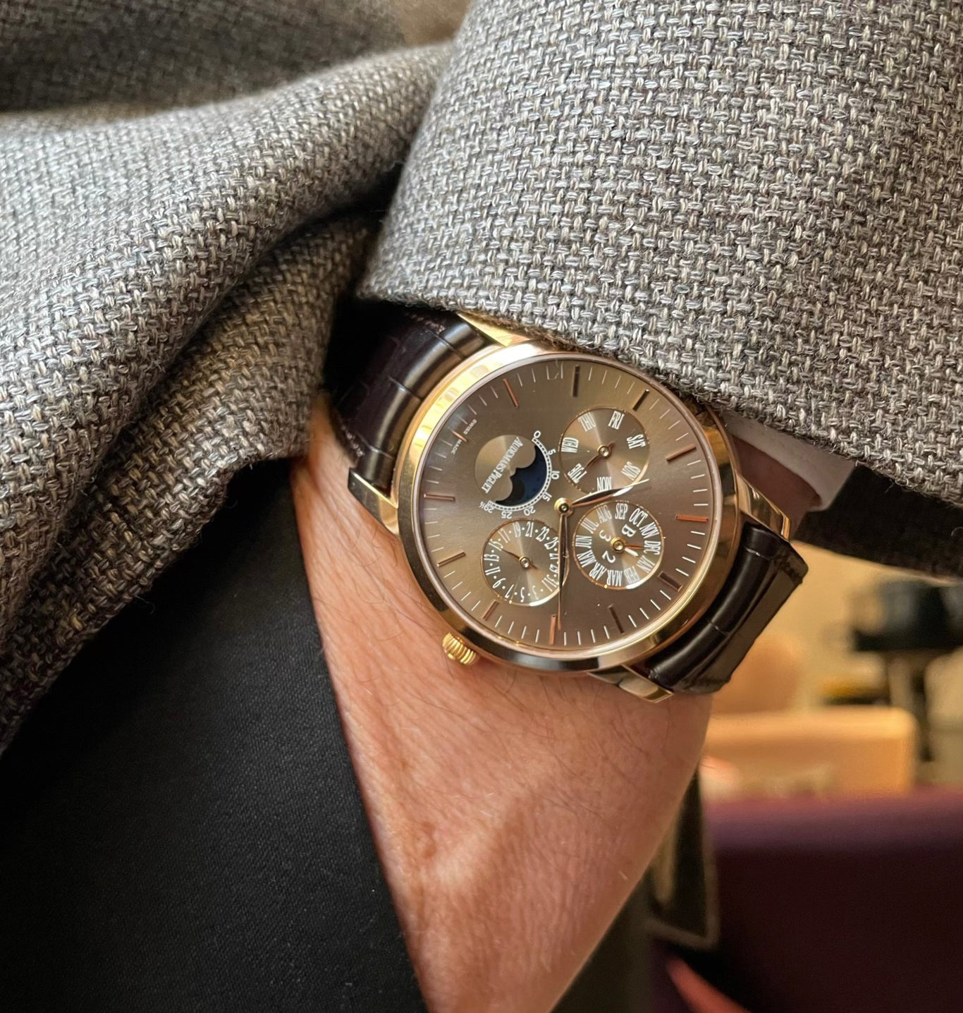A man's wrist wearing a rose gold perpetual calendar watch. He is wearing a grey textured jacket and black pants.