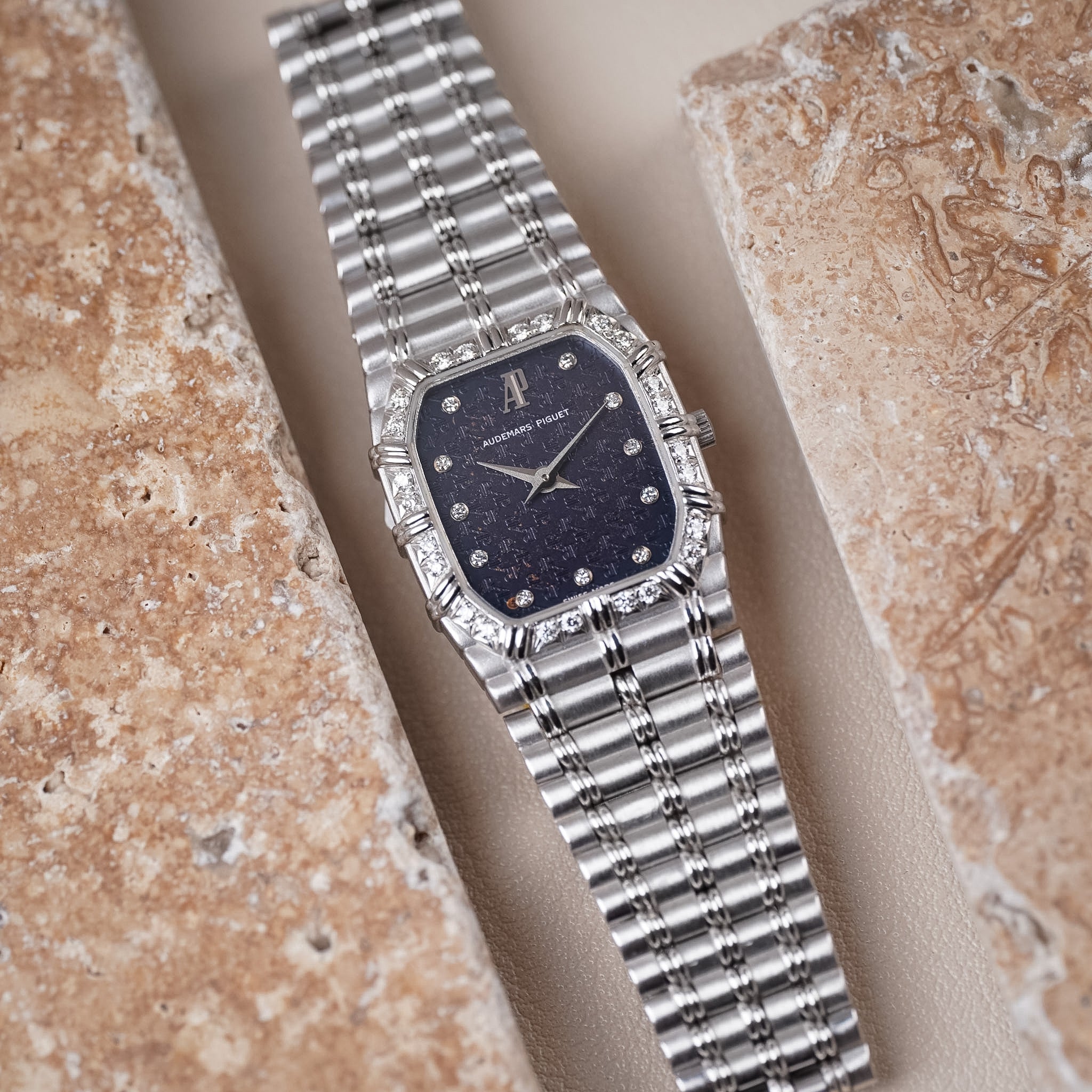 A white gold Audemars Piguet Bamboo vintage watch with a blue textured dial, diamond hour markers, white gold hands, and a diamond bezel. Shot laying flat between two beige stone tiles.