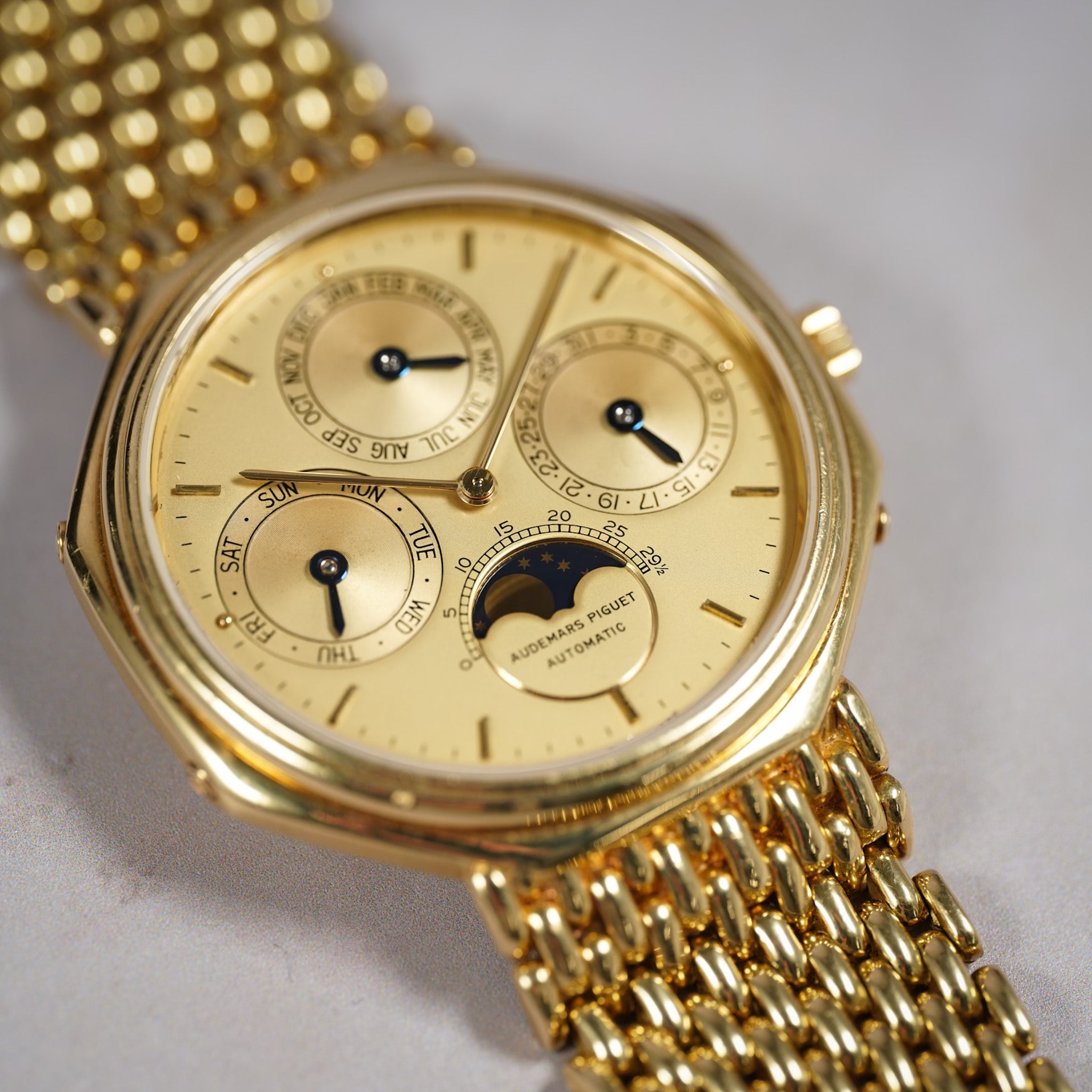 Gold-tone automatic watch with a matching gold mesh bracelet, featuring a multi-dial layout with a moonphase indicator on a light gray background.