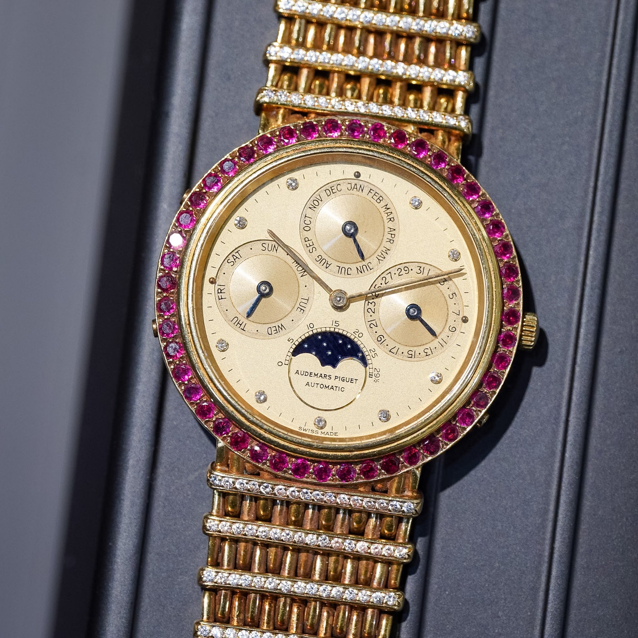A yellow gold Audemars Piguet perpetual calendar watch with a ruby bezel and four subdials laying flat on a gray tray