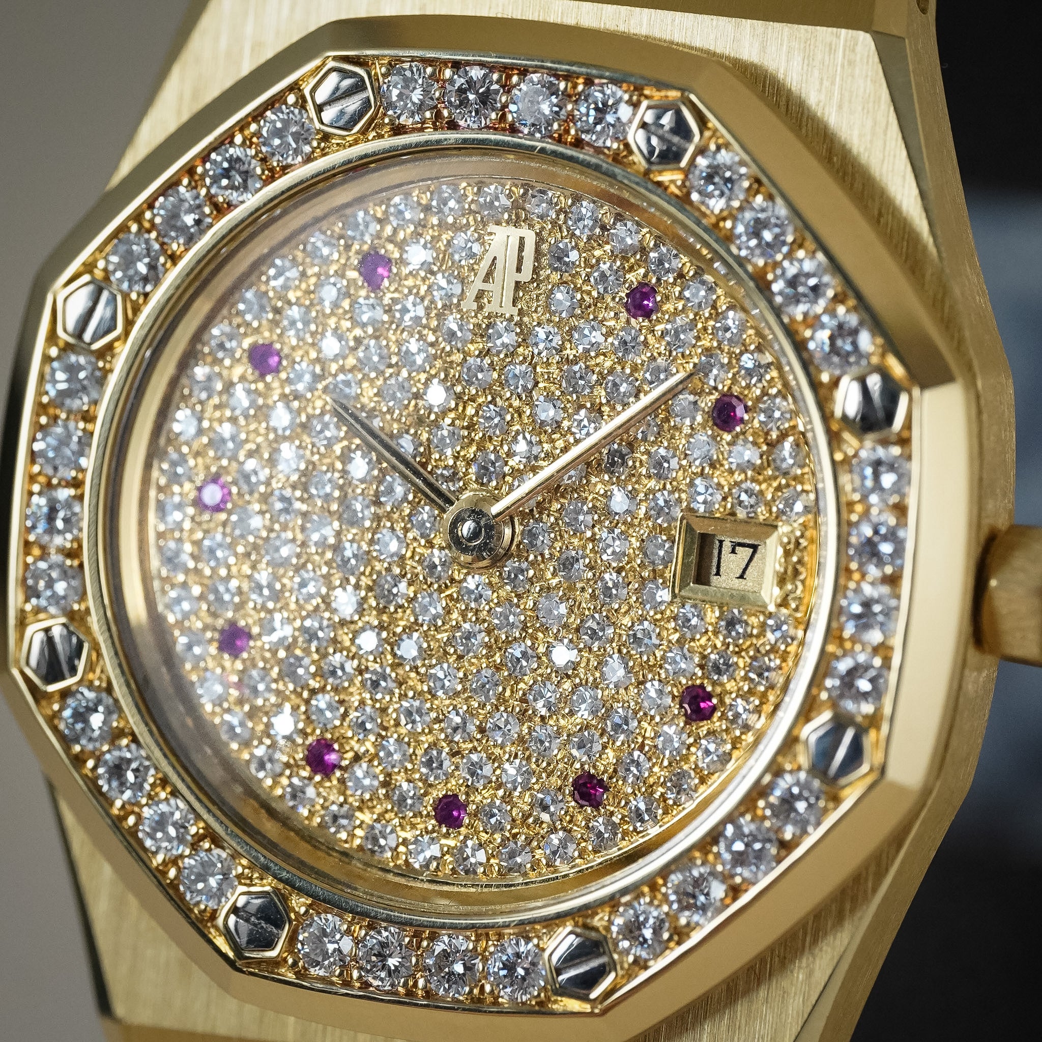Close-up of a gold-toned wristwatch with a diamond-encrusted dial, a bezel set with round stones, gold hands, and small purple accent markers on the face.
