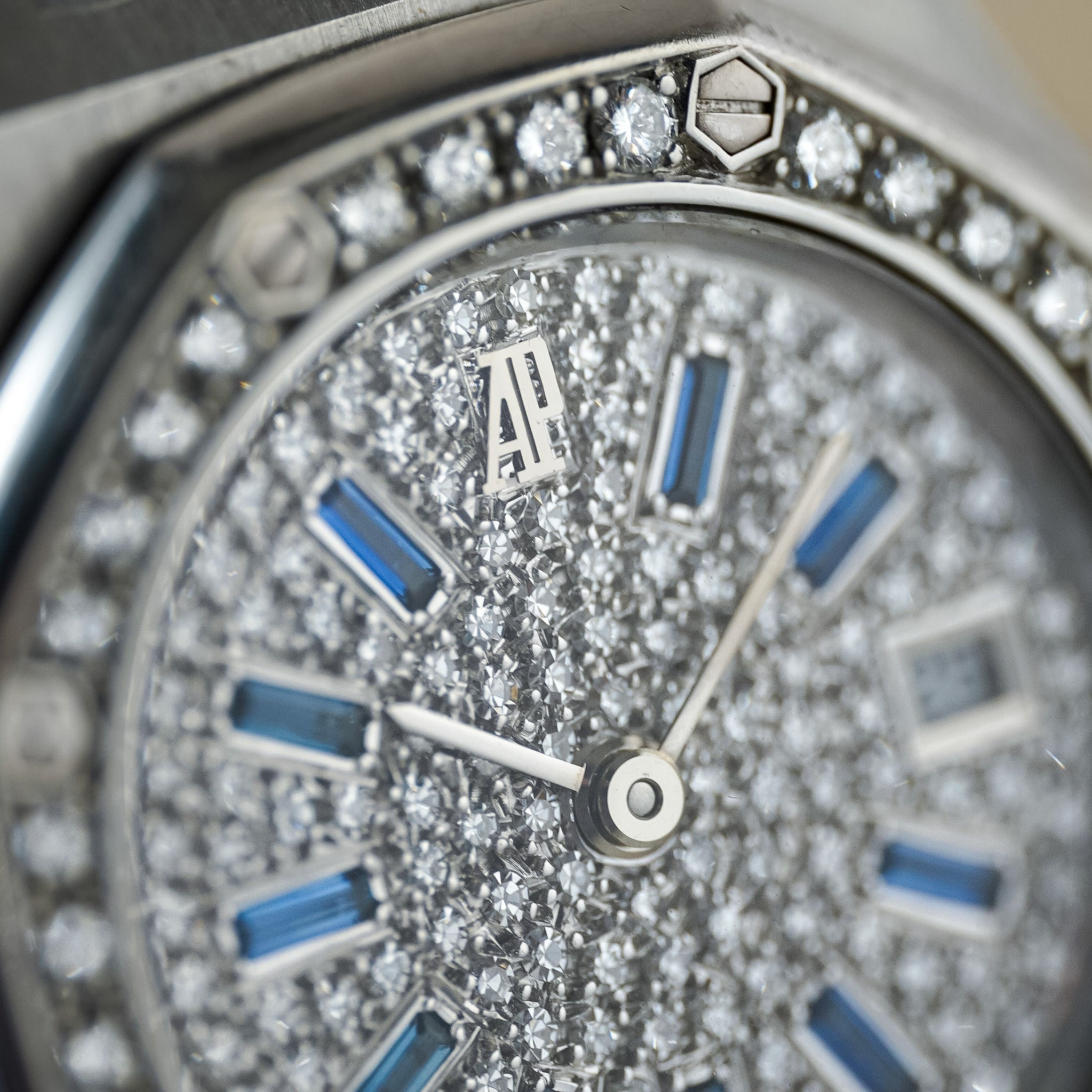 Close-up of a silver-toned wristwatch dial covered in pavé diamonds, with blue rectangular hour markers and a silver bezel accented by surrounding diamonds.