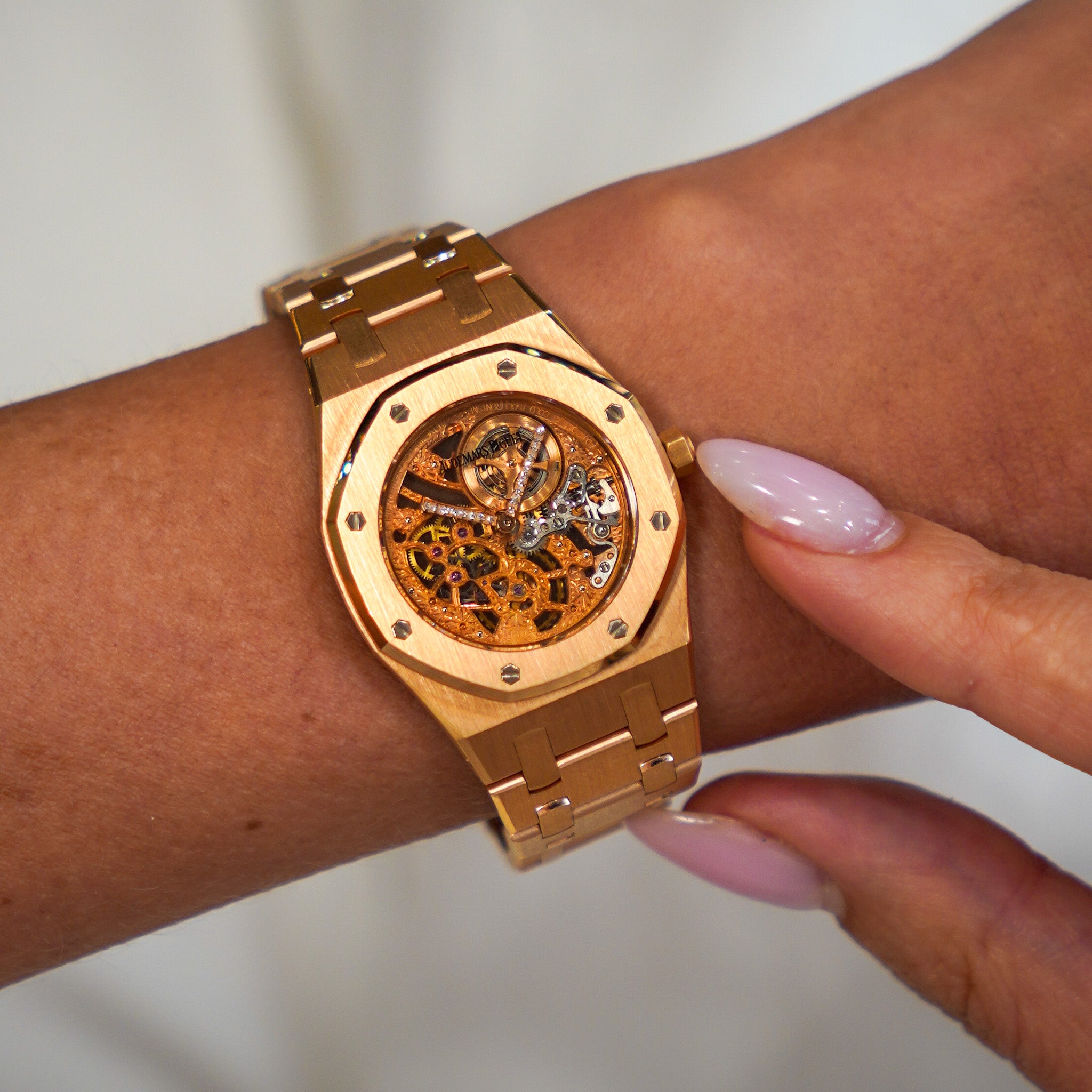 A gold-tone wristwatch with a visible skeleton dial and matching gold-tone link bracelet, shown worn on a person's wrist with a finger touching the side of the case.