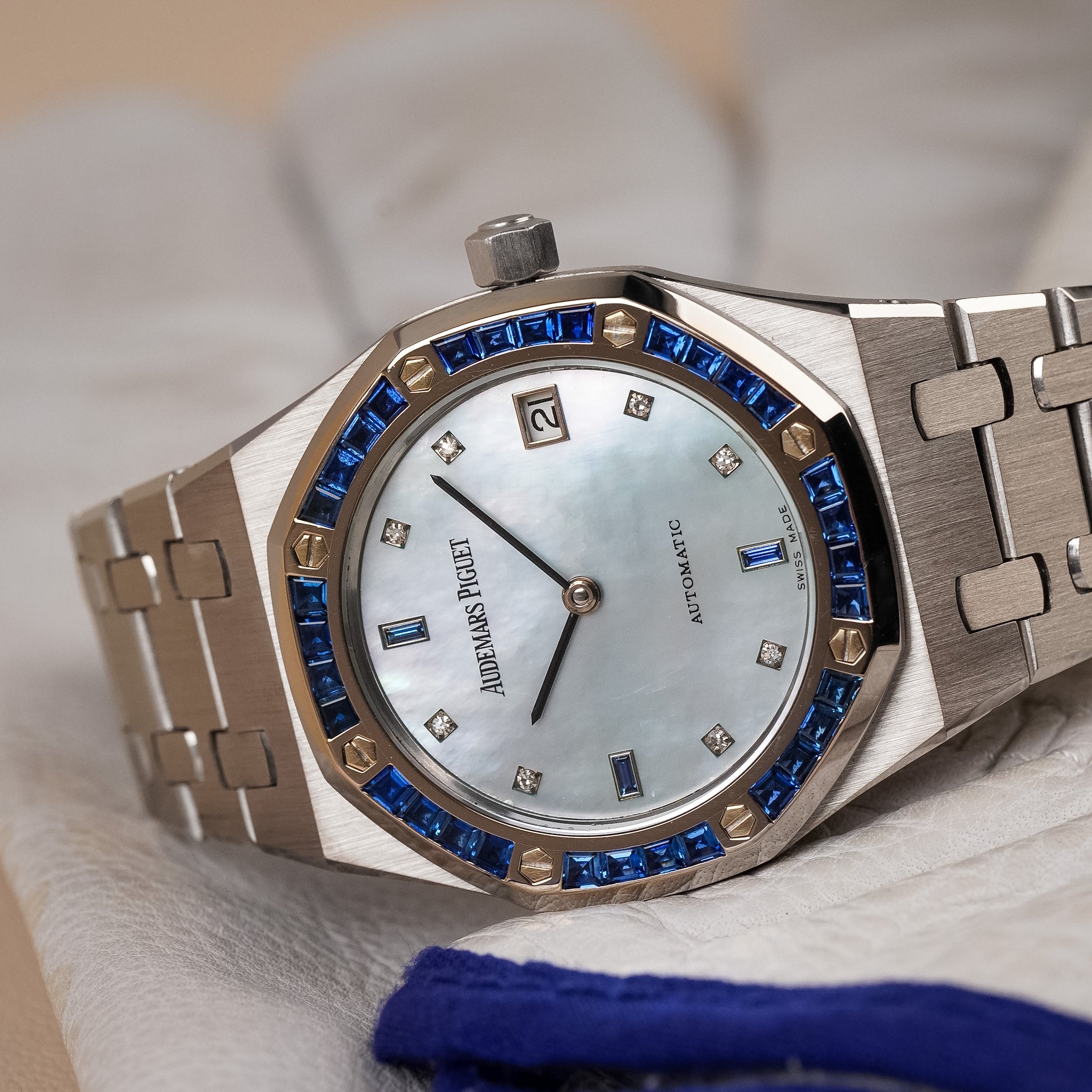 A white gold Audemars Piguet Royal Oak with a blue sapphire bezel, a blue mother of pearl dial, and blue sapphire and diamond hour markers. Shot resting sideways on a white leather surface with blue trim.