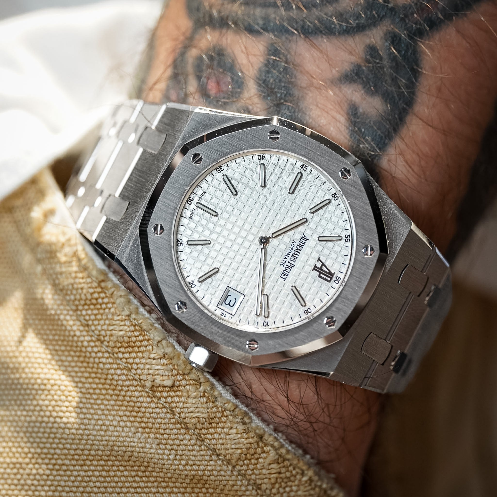 A close-up of a silver-toned stainless steel wristwatch with a white textured dial, worn on a person's tattooed wrist.