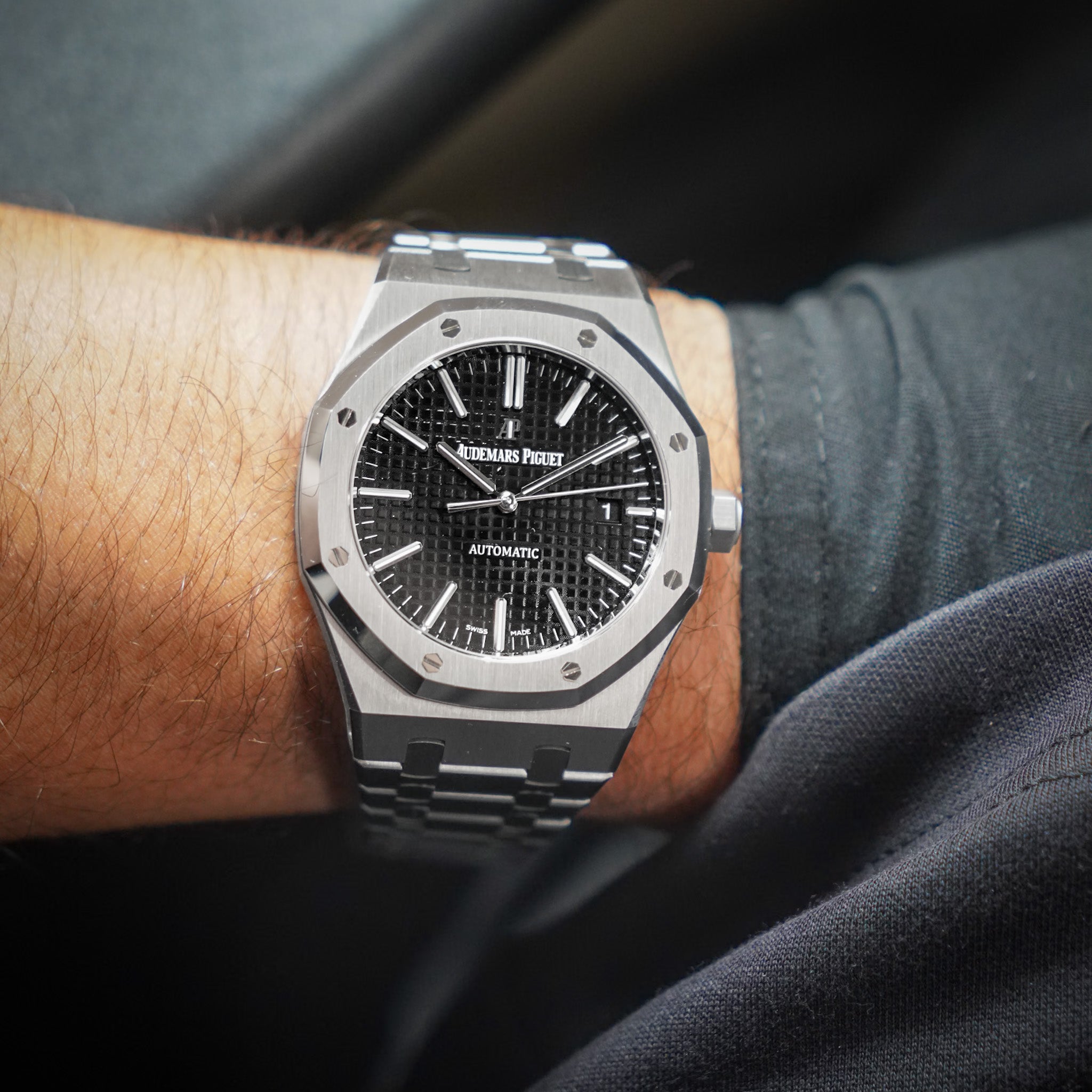 A close-up shot of a wrist wearing an Audor Automatic watch with a silver-toned octagonal bezel, black textured dial, and matching metal link bracelet.