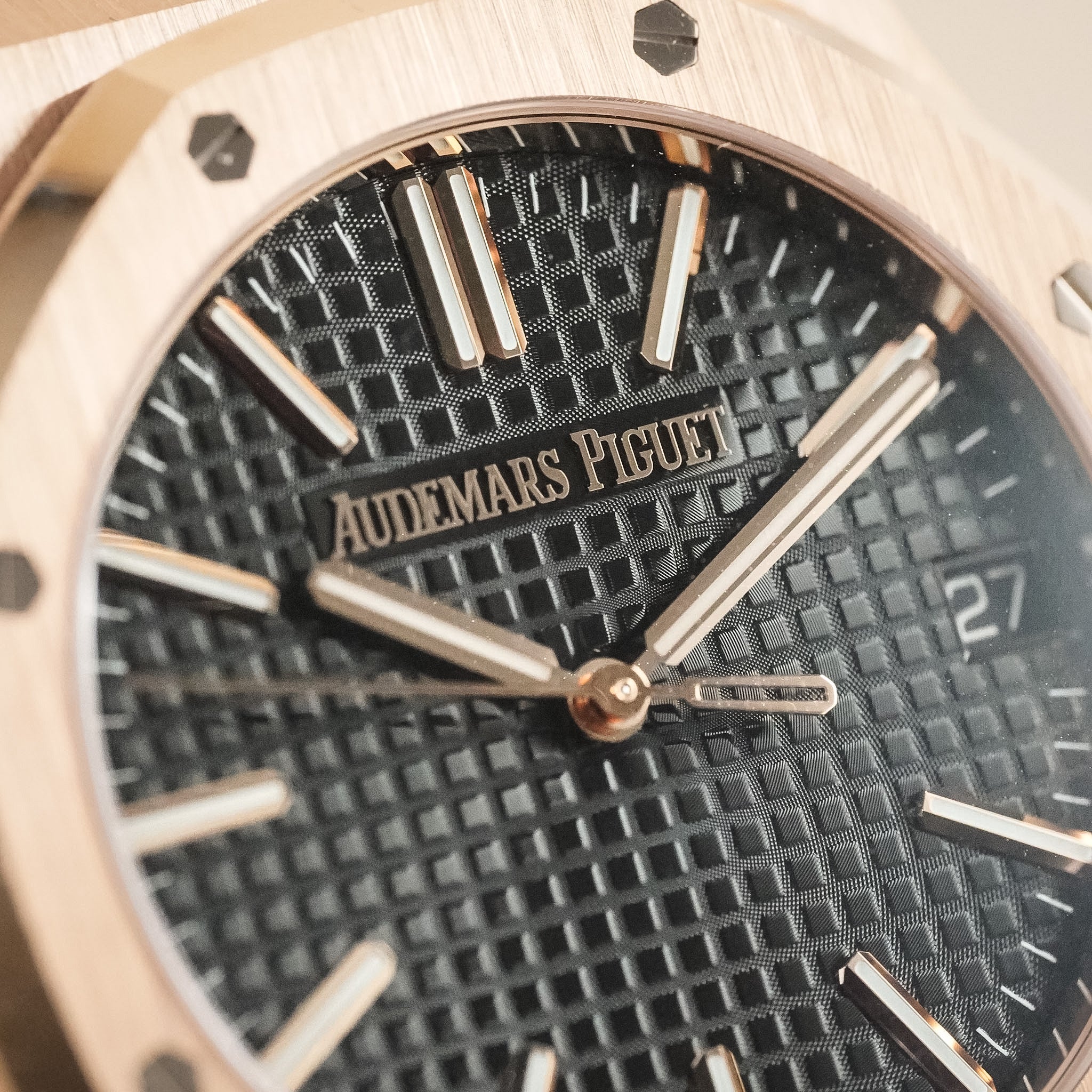 A closeup of a textured black watch dial. The brand name Audemars Piguet is visible, as is a date window showing the number 27. The hands, bezel, and hour markers are in rose gold