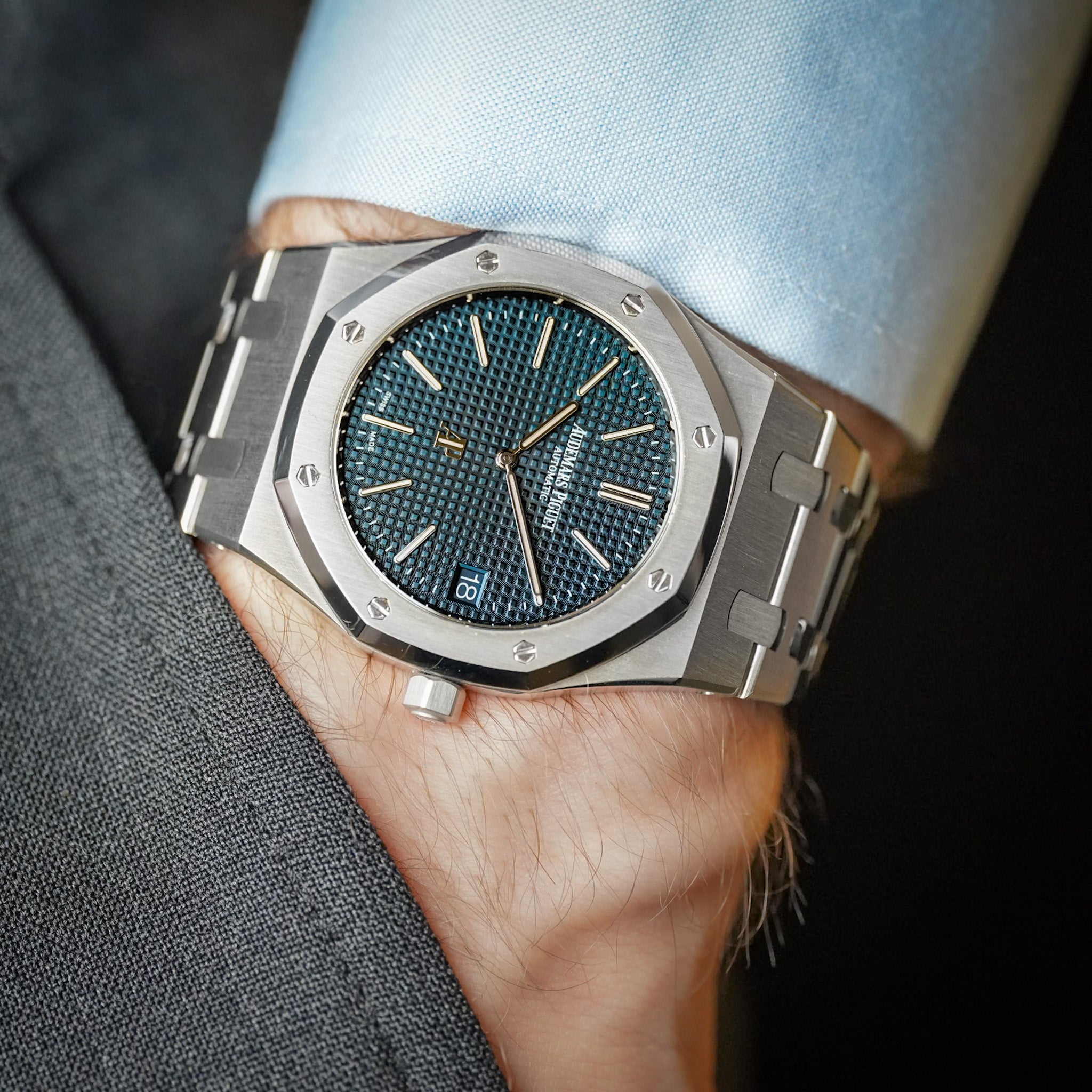 A steel Audemars Piguet Royal Oak with a textured blue dial, octagonal bezel with exposed screws, and a date window showing "18", shot on a man's wrist waring a blue shirt and grey pants