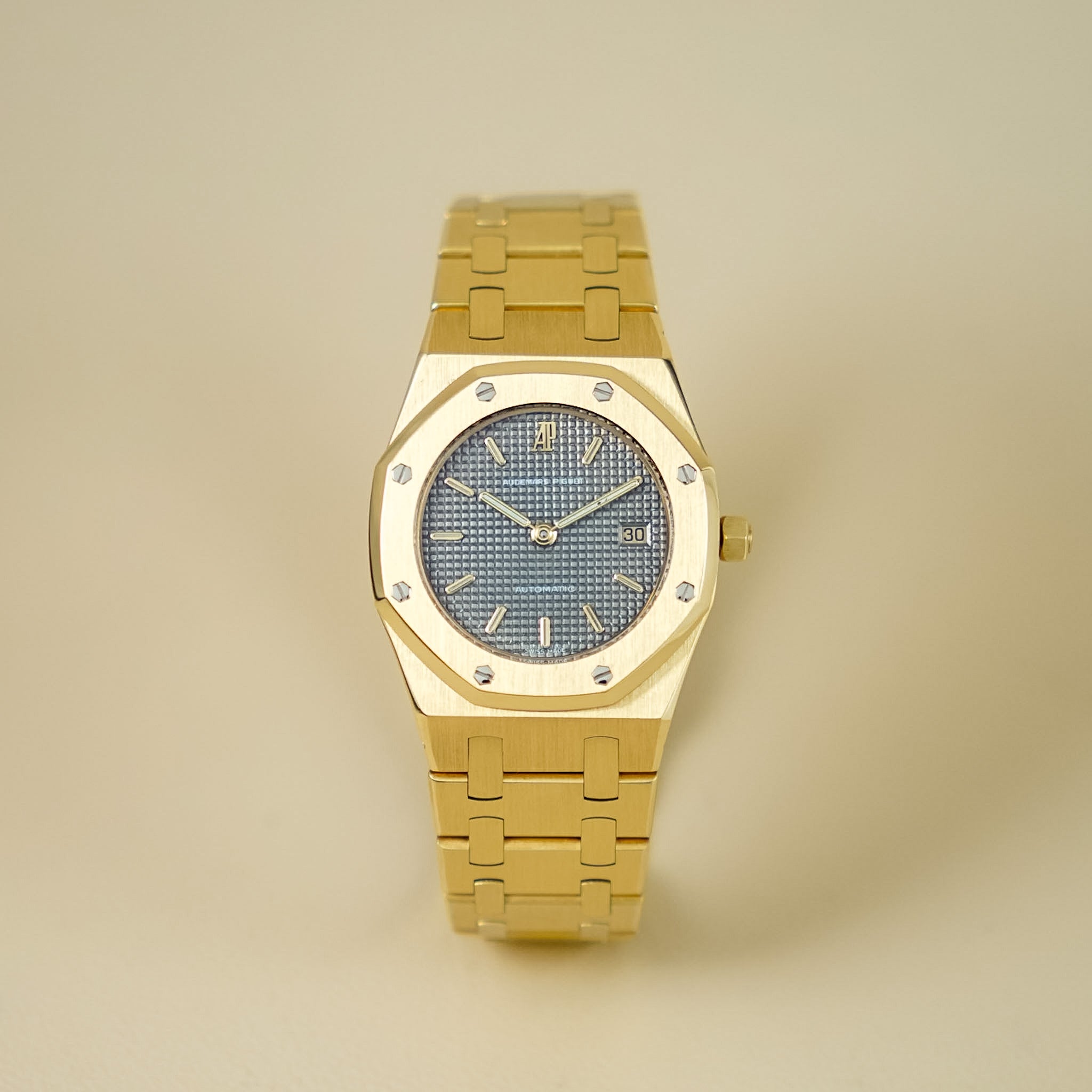 An Audemars Piguet Royal Oak ladies watch with a full yellow gold case and bracelet, grey dial, shot against a neutral cream background
