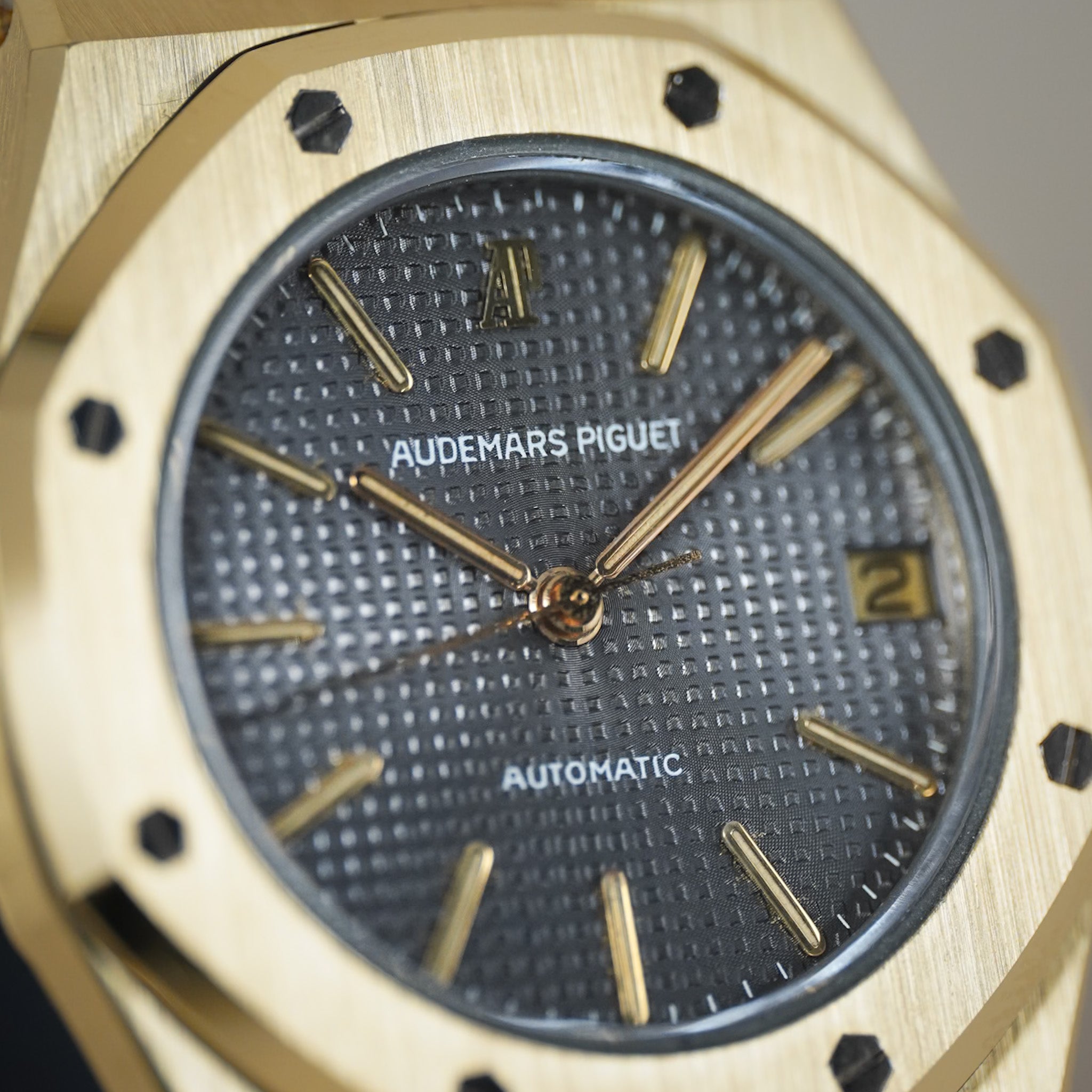 Close-up of an Audemars Piguet Automatic watch with a brushed gold-tone case, textured dark gray dial, and gold-tone hour markers and hands.