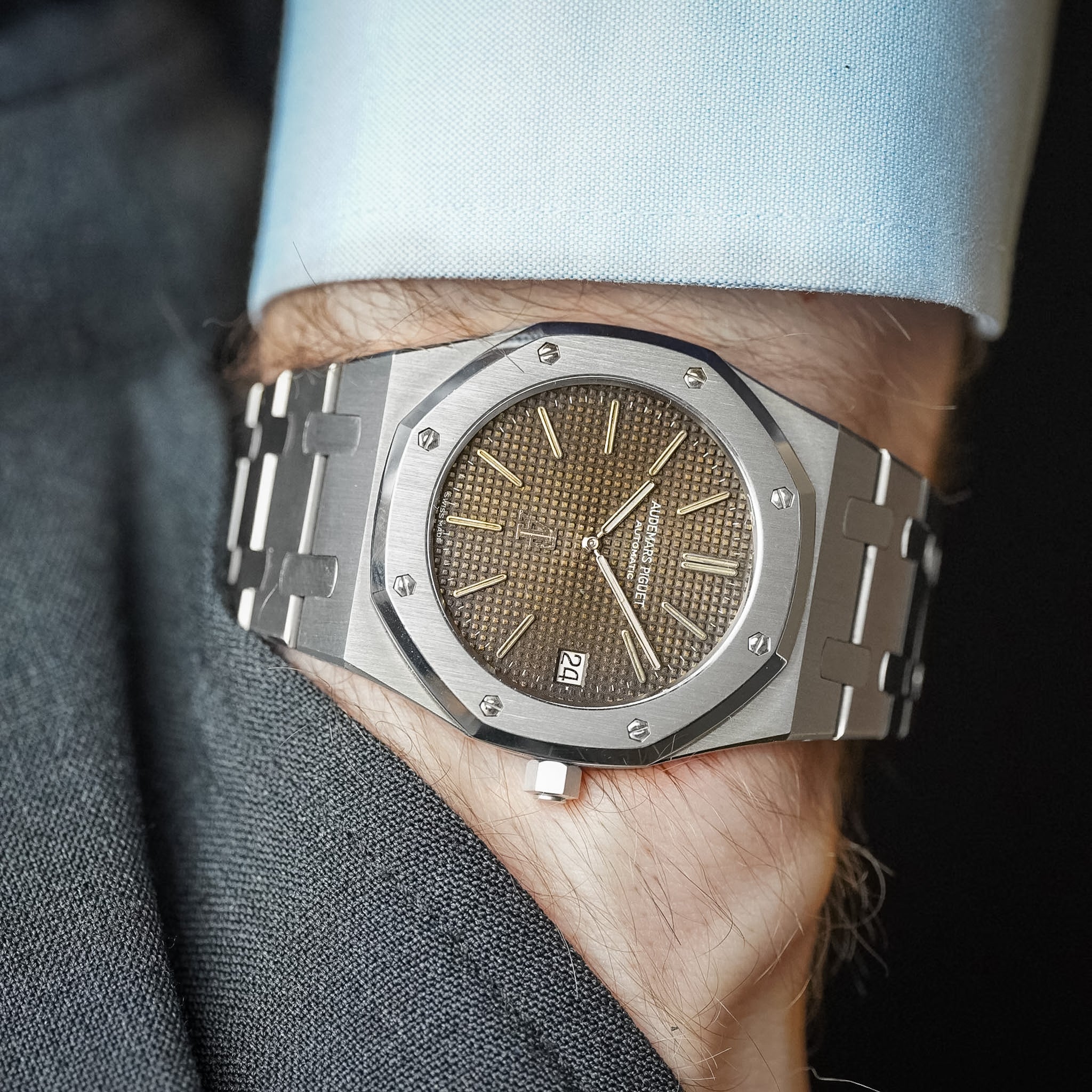 An Audemars Piguet Royal Oak in steel with a tropical brown dial, octagonal bezel with exposed screws, and a stainless steel bracelet, shot on a man's wrist wearing a light blue shirt and a grey pants