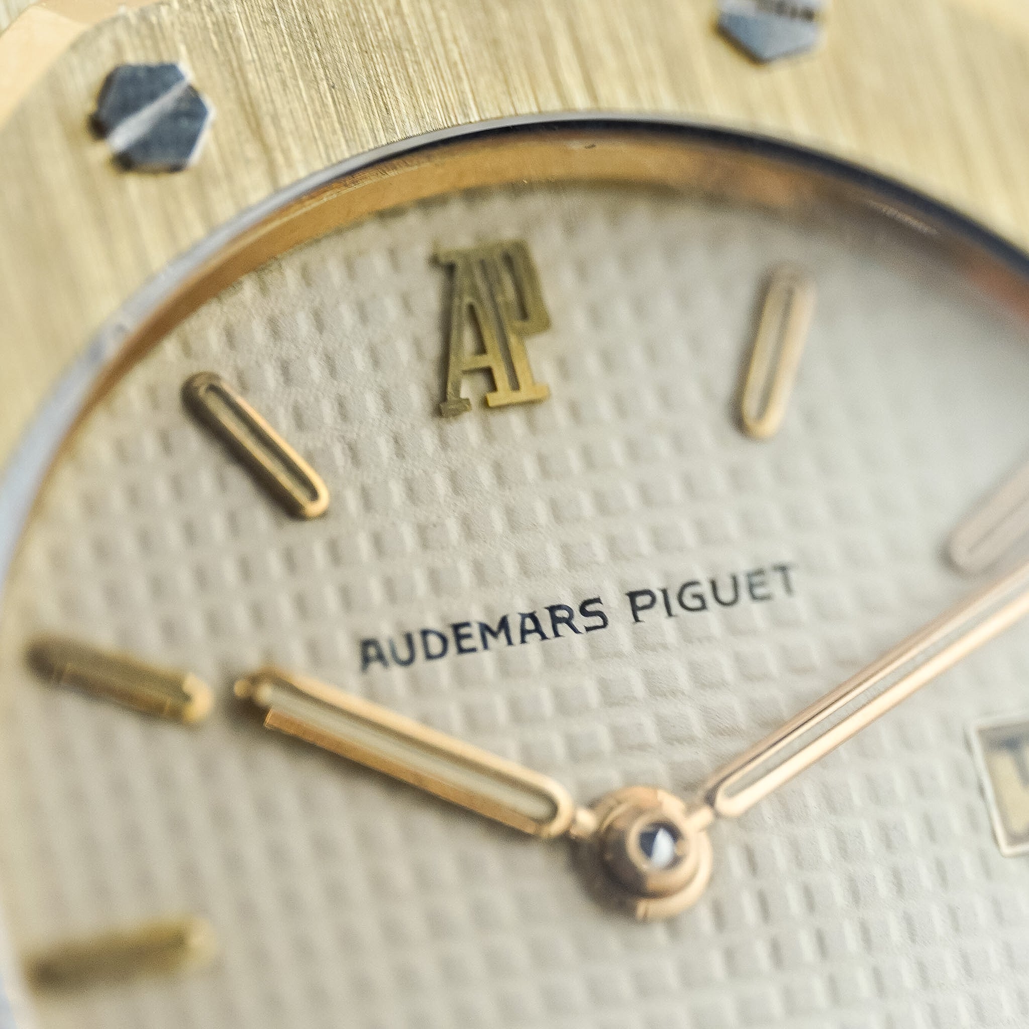 Close-up of an Audemars Piguet watch dial and bezel, showing a textured champagne dial with gold-tone hands, indices, and logo, and a brushed gold-tone case.