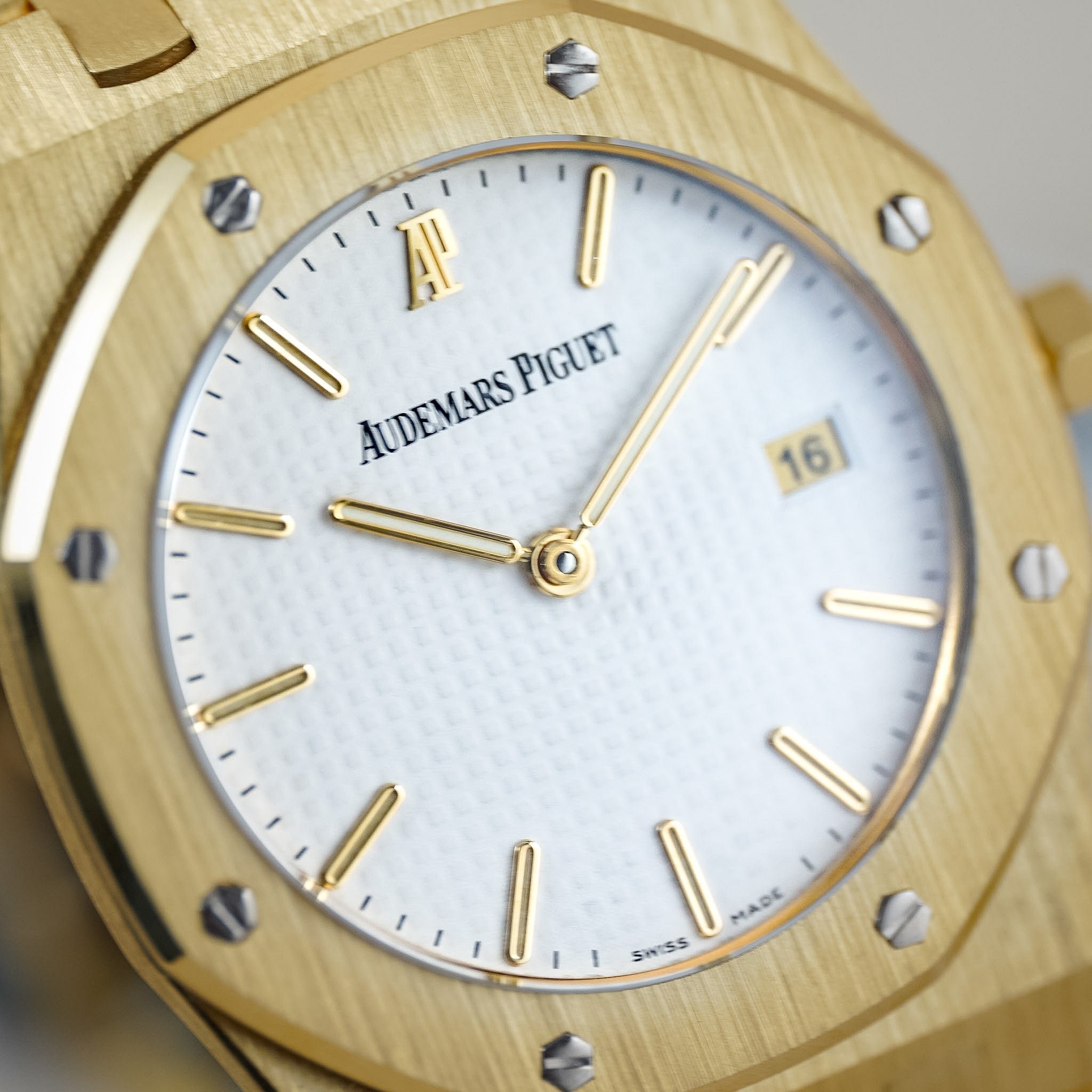 Close-up of an Audemars Piguet Royal Oak watch with a brushed gold-tone case, white dial, gold-tone hour markers and hands, and a date window at 3 o'clock.